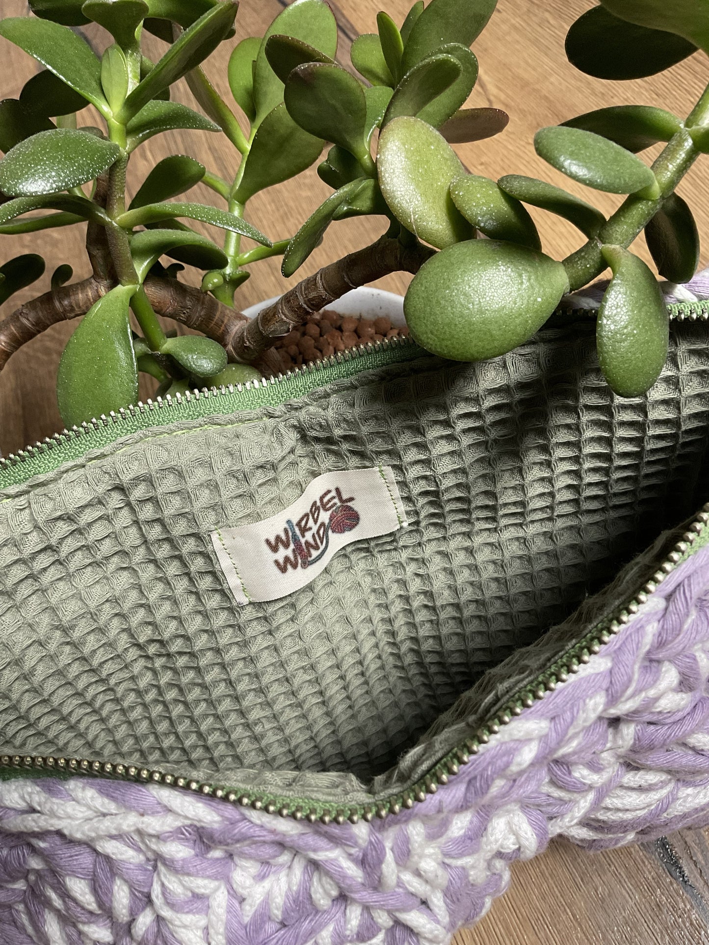 Der Reißverschluss der Tasche ist geöffnet, wir sehen das salbeigrüne Innenfutter aus Waffelstoff. Die Tasche steht neben einer Sukkulente.