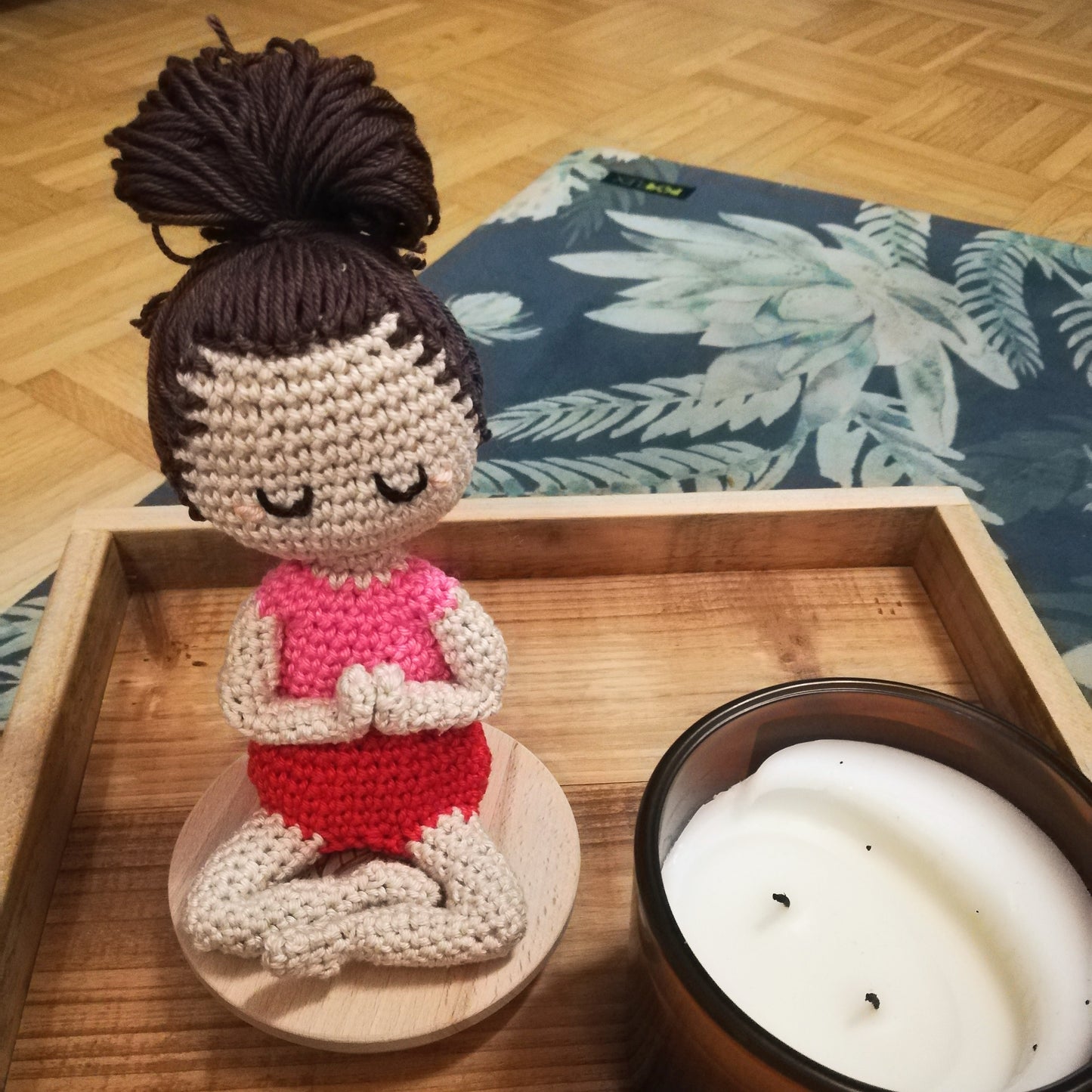 Weibliche Puppe, heller Hautton, Hände vor dem Herzen zusammen, im Schneidersitz, Augen geschlossen, rosa Wangen, dunkelbrauner großer Dutt, pinkes Top, rote Shorts. Neben ihr eine Kerze, beides steht auf einem Holztablett auf einer grünen Yogamatte mit hellgrünen Blumen.