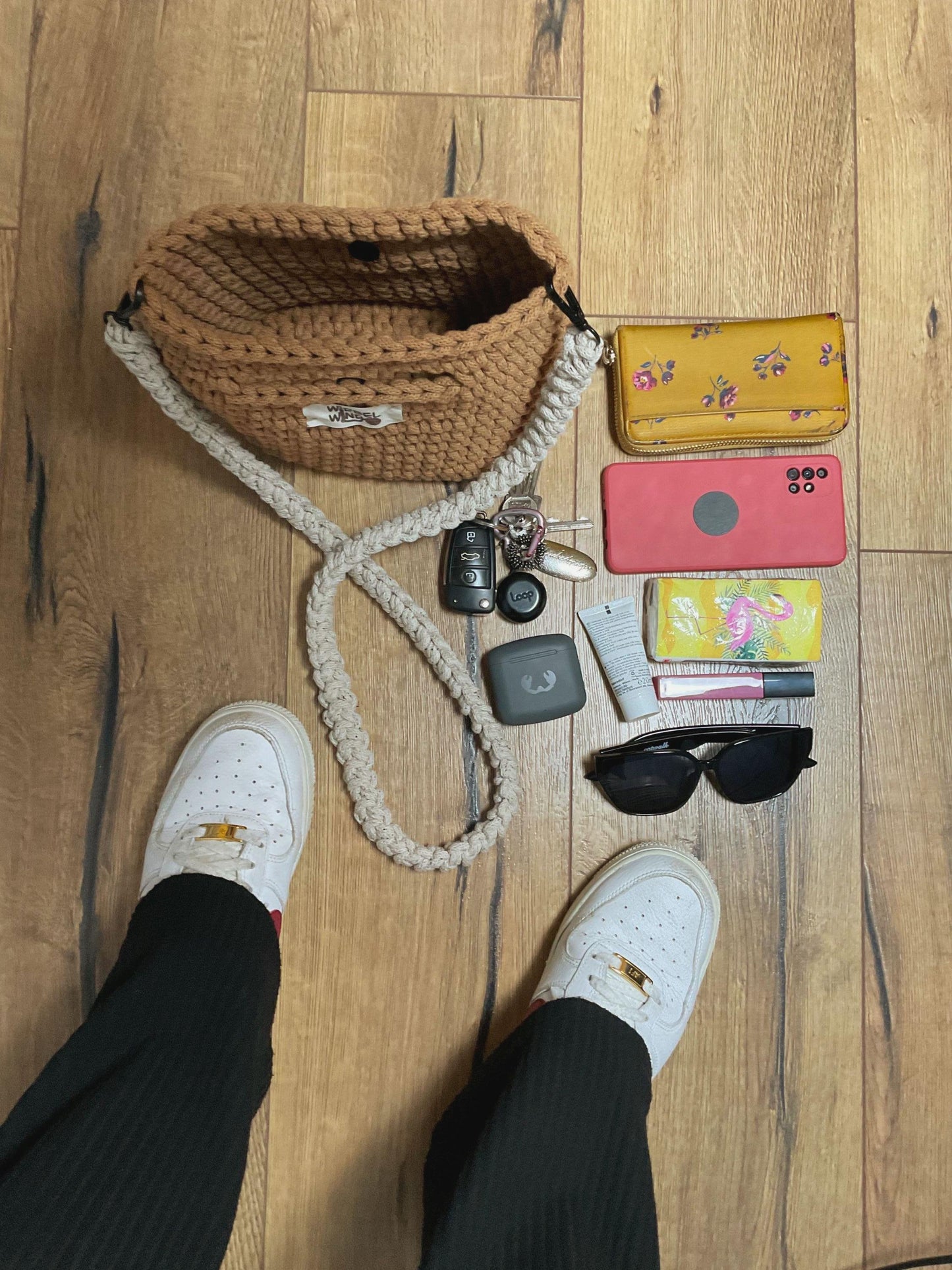 Die Tasche ist geöffnet und steht auf dem Boden, Blick von oben. Daneben liegen: Schlüssel, Handy, Geldbeutel, Kopfhörer, Taschentücher, Lippenstift, Handcreme, Sonnenbrille. Eine Person steht daneben, wir sehen die weißen AF1 Nike Schueh und schwarze Schlaghosen.