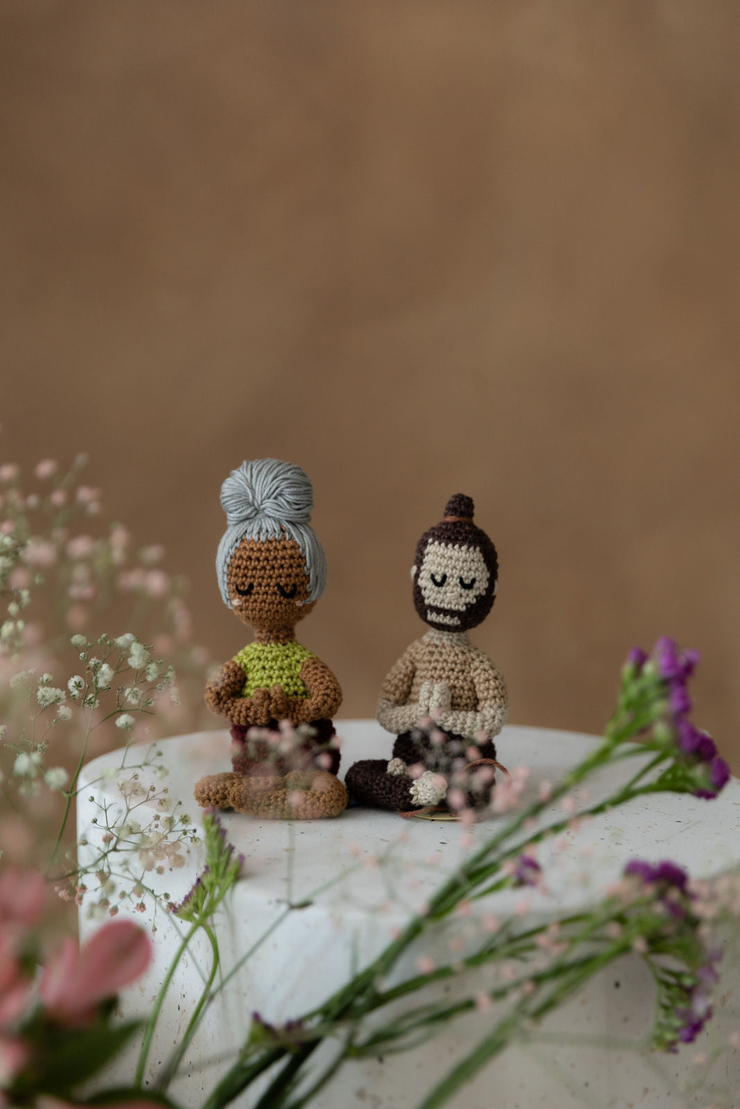 Weibliche und männliche Puppe von weitzer weg fotografiert, sitzen auf weißwm Bettonklotz, Hintergrund braun, vor ihnen Blumen in rosa, weiß, lila, pink, alle unscharf gestellt.