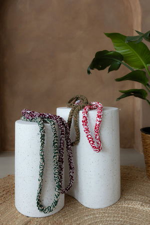 Zwei kleine Betonsäulen, darauf 4 gehäkelte Lanyards. Hintergrund braun-limewash Wand mit Bogen, runder Juteteppich, große grüne Pflanze ragt rechts ins Bild.