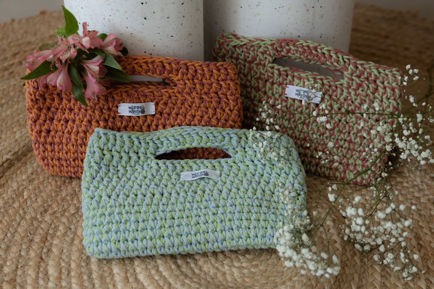 2 Clutches hinten, eine vorne , alle überlappen sich und sind gegen 2 Betonsäulen gelehnt. Alles steht auf Juteteppich. Rechts ragt weißes Schleierkraut ins Bild. Auf der Tasche links oben liegen Blumen mit rosa Blüten.