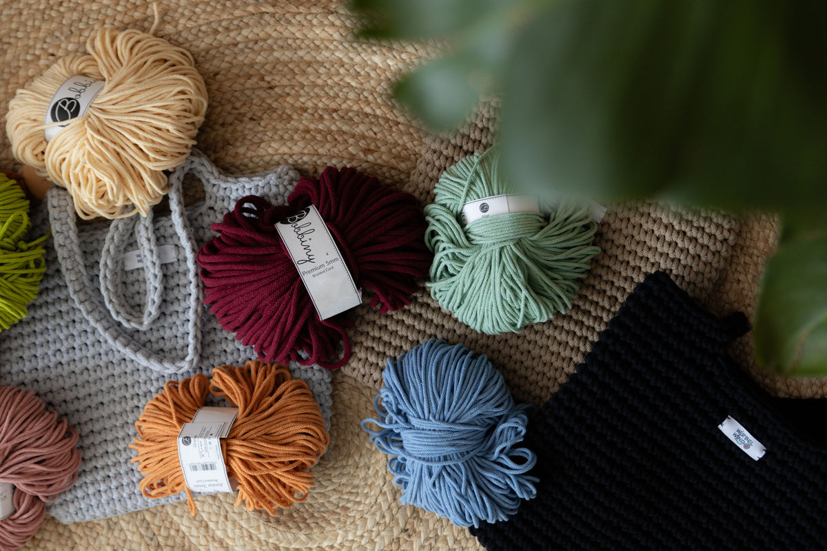 Jute-Teppich auf dem Boden. 3 Shopper in grau, beige, schwarz liegen auf dem Boden. Bunte Wollknäule in rot, blau, grün, orange, rosa, gelb liegen darauf verstreut. Von rechts ragt verschwommen grüne Pflanze ins Bild.