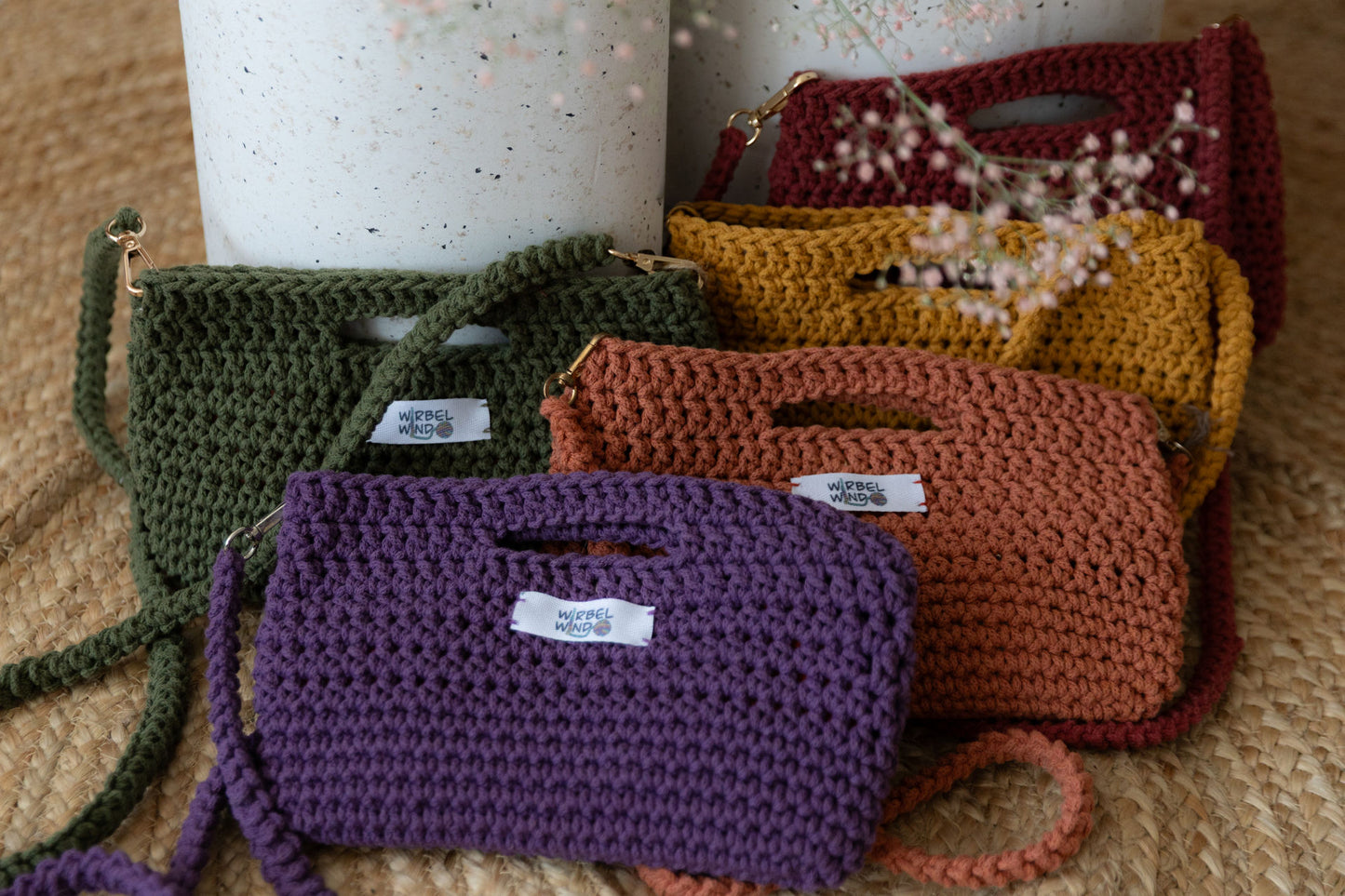 5 gehäkelte Molly Mini Taschen in Violet, Orange, Grün, Gelb, Rot gegen zwei runde Betonsäulen gelehnt. Von oben ragt rosa Schleierkraut ins Bild, ist aber unscharf.