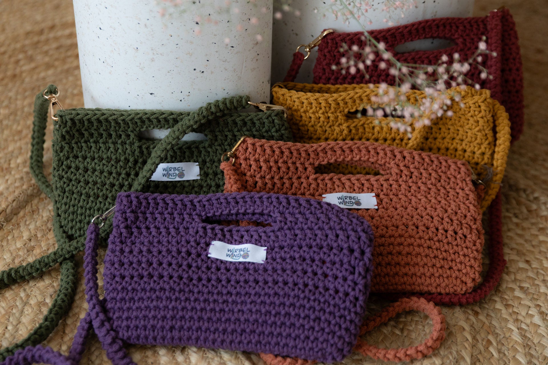 5 gehäkelte Molly Mini Taschen in Violet, Orange, Grün, Gelb, Rot gegen zwei runde Betonsäulen gelehnt. Von oben ragt rosa Schleierkraut ins Bild, ist aber unscharf.