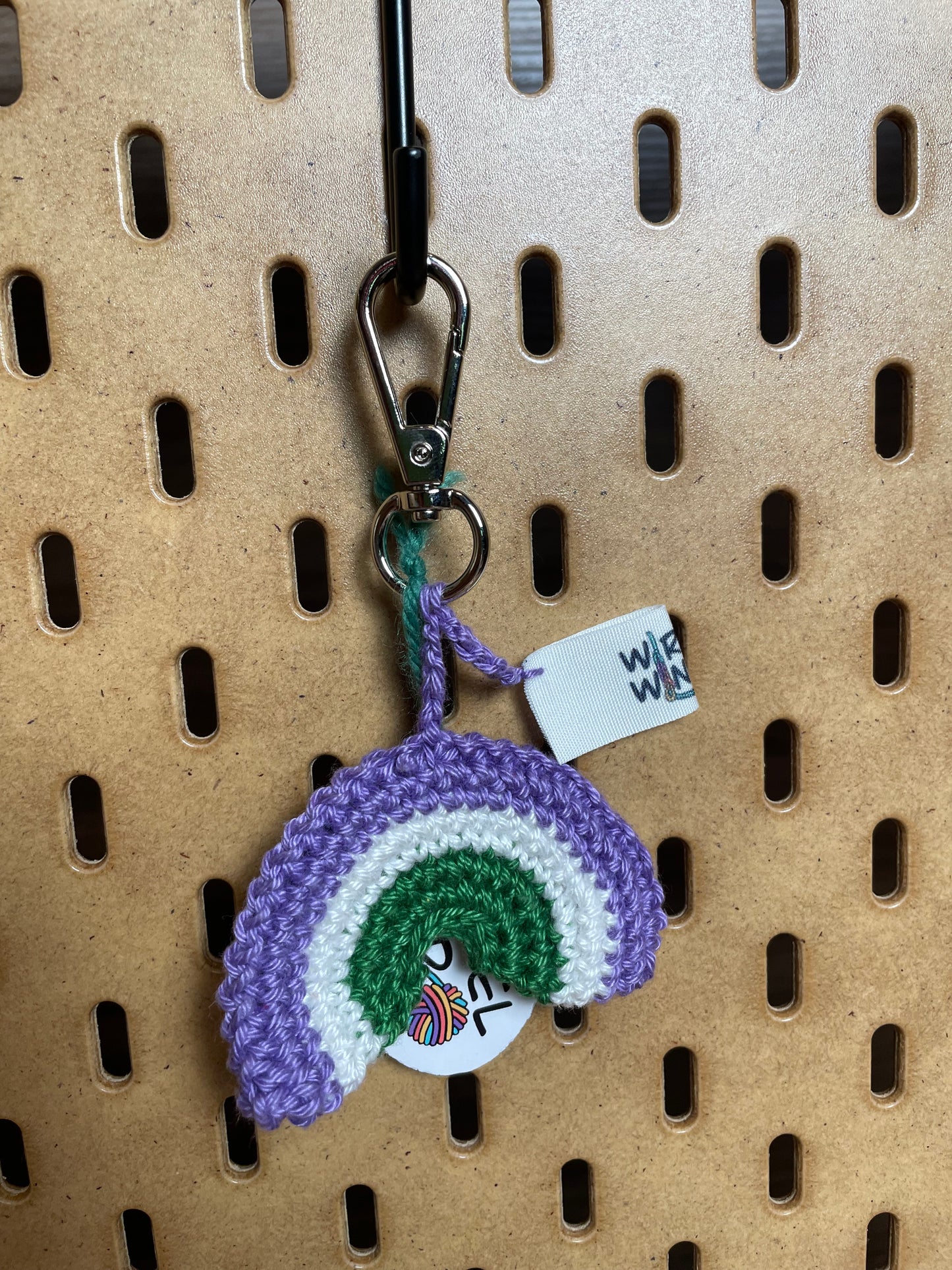 Gehäkelter Regenbogen Schlüsselanhänger mit Karabiner in den Farben der Genderqueer/Nonbinary Pride Flagge: Lila, Weiß, Grün. Außerdem hängt das Logo von Wirbelwind ebenfalls am Karabiner.