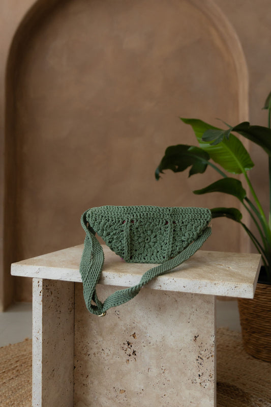 Gehäkelte Tasche in Eukalyptusrün in Bananenform mit verstellbarem Umhängegurt steht auf einem kleinen Tisch aus Stein in Beigetönen. Im Hintergrund eine braune Limewash-Wand mit einer Bogen-Nische. Auf dem Boden ein runder Teppich aus Jute, sowie eine Pflanze mit großen grünen Blättern in einem hellbraunen Topf, das Material ähnlich Bambus.