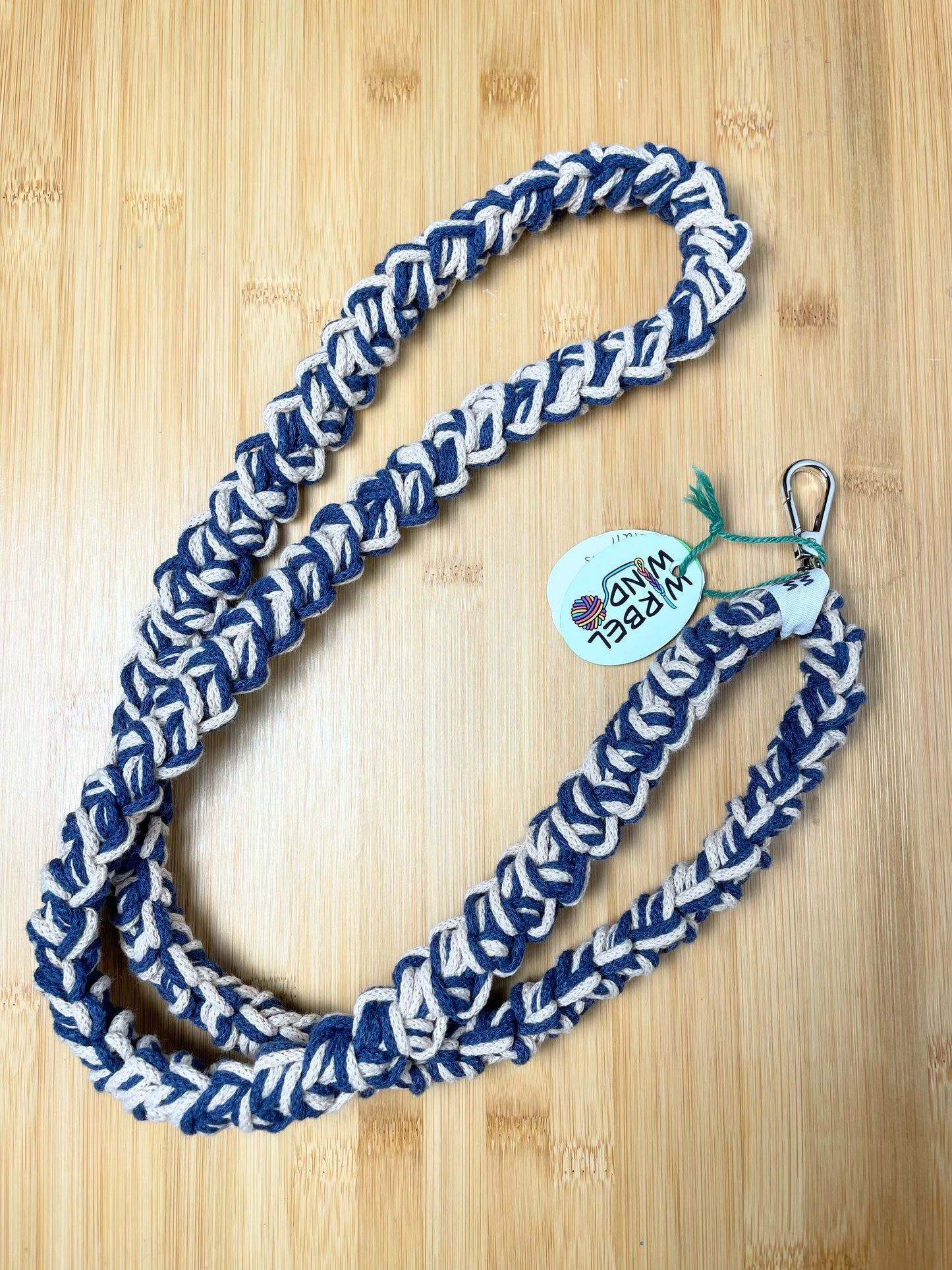 Beige-Jeansblau-meliertes Lanyard kringelt sich. Wirbelwindlogo am unteren Ende, direkt über silbernem Karabiner angenäht.