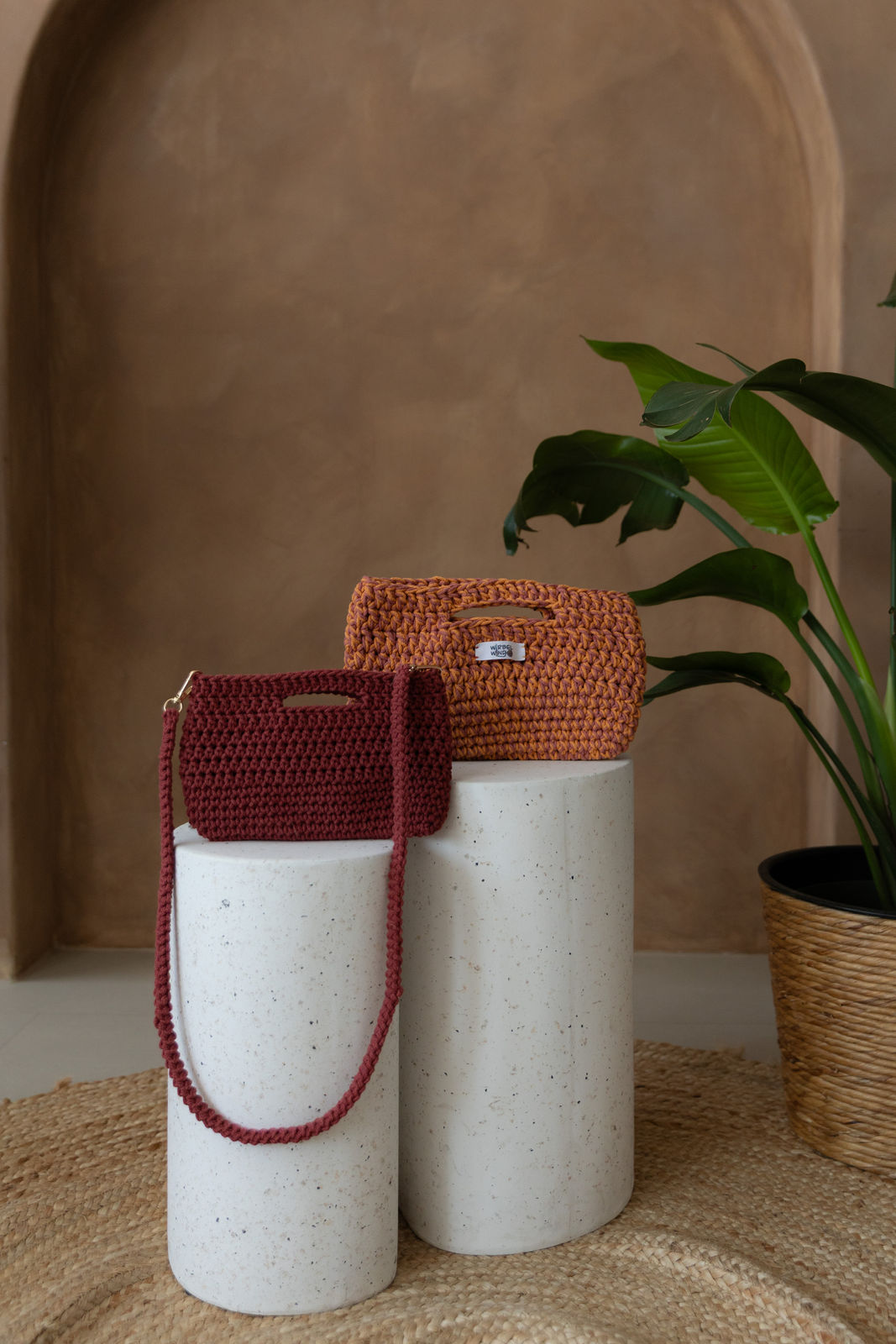 2 unterschiedlich hohe runde Betonsäulen. Auf ihnen rechts und etwas nach hinten versetzt eine Molly Medium Tasche in Pink & Orange, davor und links eine Molly Mini Tasche in Weinrot, ihr Umhängegurt hängt vorne nach unten. Rechts ragt grüne Pflanze ins Bild. Hintergrund ist braune Limewash Wand mit Bogen.