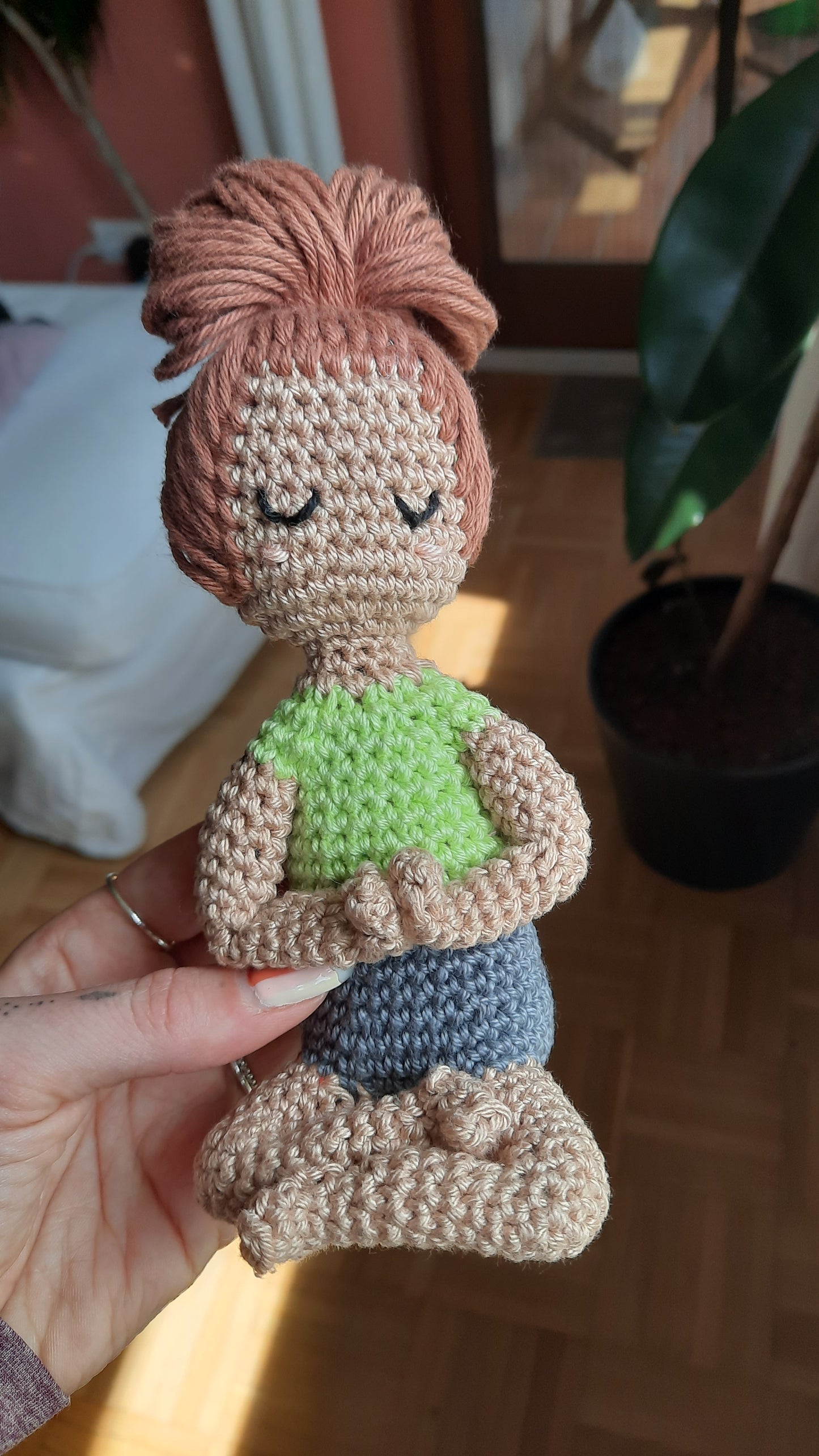 Weibliche Puppe, Hände vor dem Herzen zusammen, Schneidersitz, helle Haut, hellbraune Haare in großem Dutt, hellgrünes, ärmelloses Shirt, graue Shorts, Augen geschlossen, pfirsichfarbene Wangen.
