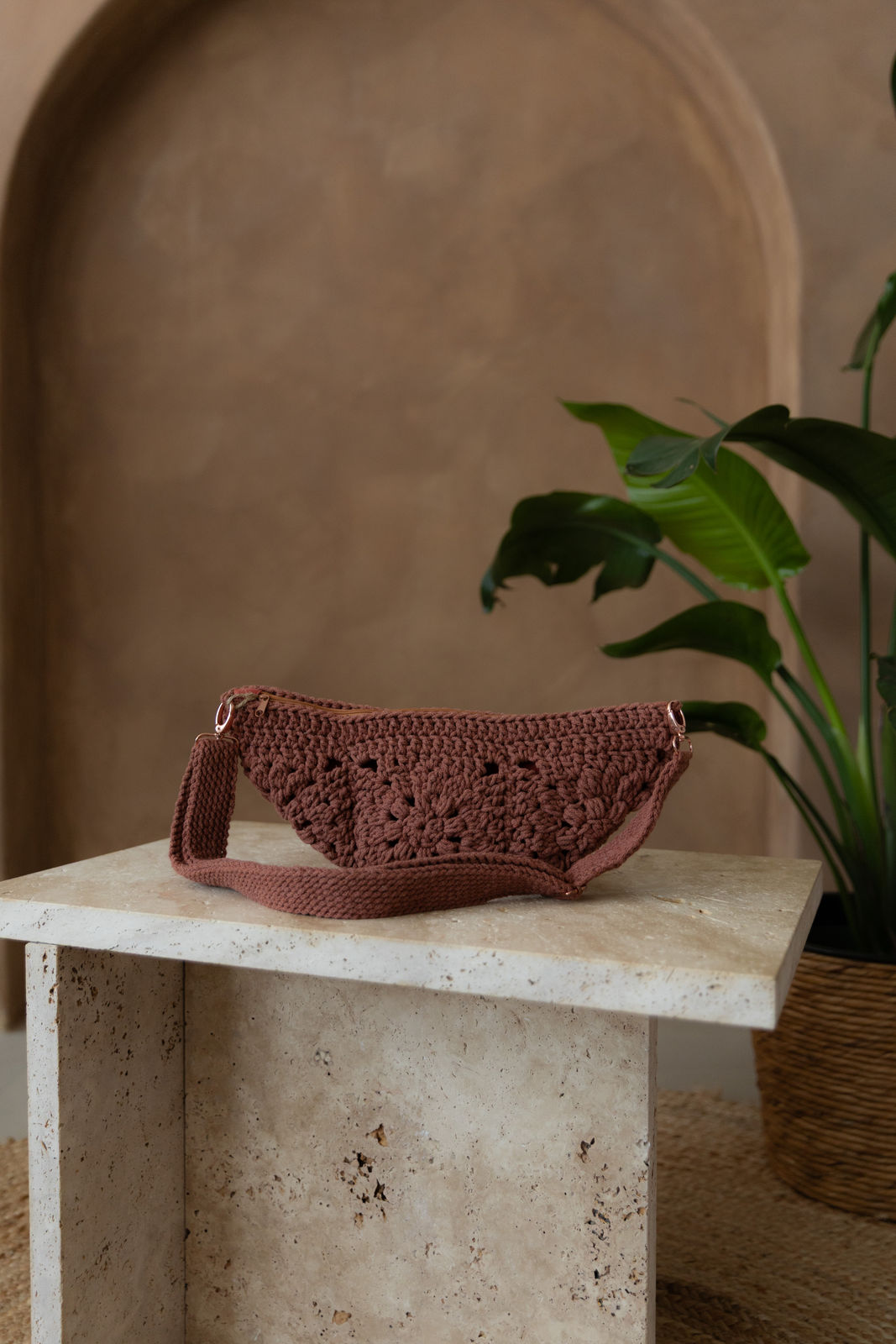 Gehäkelte Tasche in der Farbe Sunset  in Bananenform mit verstellbarem Umhängegurt steht auf einem kleinen Tisch aus Stein in Beigetönen. Im Hintergrund eine braune Limewash-Wand mit einer Bogen-Nische. Auf dem Boden ein runder Teppich aus Jute, sowie eine Pflanze mit großen grünen Blättern in einem hellbraunen Topf, das Material ähnlich Bambus.