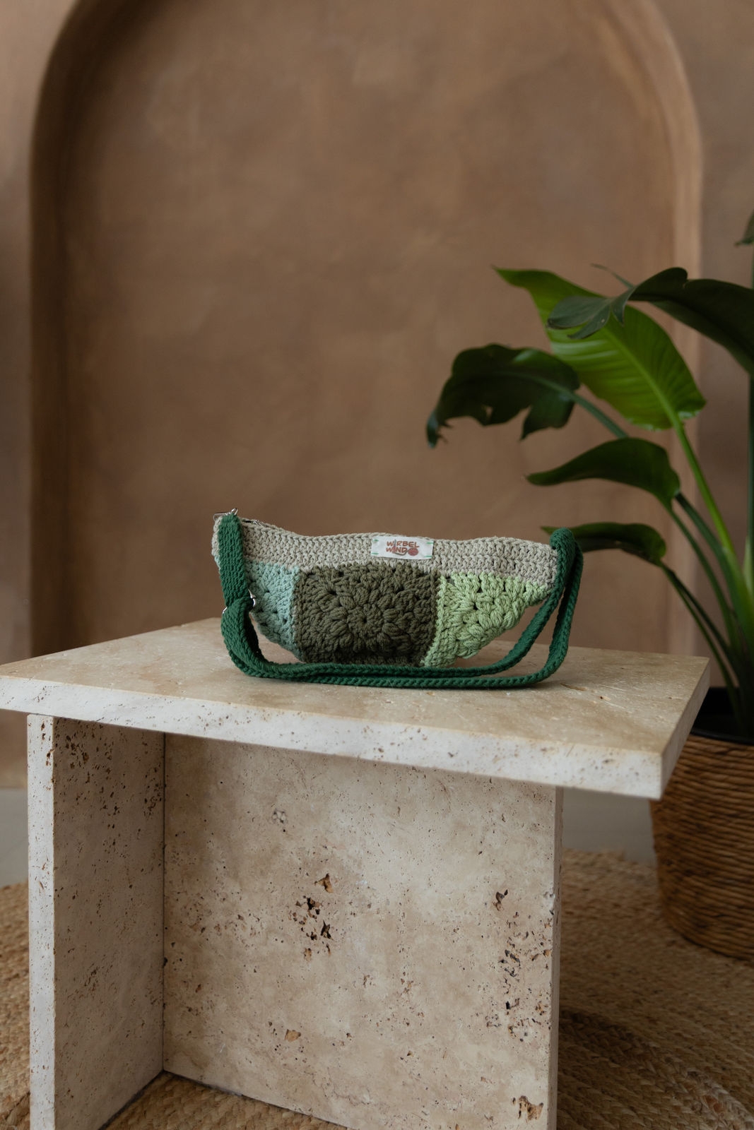 Gehäkelte Tasche in 10 verrschiedenen Gröntönen in Bananenform mit verstellbarem Umhängegurt steht auf einem kleinen Tisch aus Stein in Beigetönen. Im Hintergrund eine braune Limewash-Wand mit einer Bogen-Nische. Auf dem Boden ein runder Teppich aus Jute, sowie eine Pflanze mit großen grünen Blättern in einem hellbraunen Topf, das Material ähnlich Bambus.