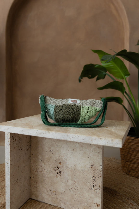 Gehäkelte Tasche in 10 verrschiedenen Gröntönen in Bananenform mit verstellbarem Umhängegurt steht auf einem kleinen Tisch aus Stein in Beigetönen. Im Hintergrund eine braune Limewash-Wand mit einer Bogen-Nische. Auf dem Boden ein runder Teppich aus Jute, sowie eine Pflanze mit großen grünen Blättern in einem hellbraunen Topf, das Material ähnlich Bambus.