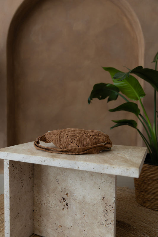 Gehäkelte Tasche in Karamellbraun in Bananenform mit verstellbarem Umhängegurt steht auf einem kleinen Tisch aus Stein in Beigetönen. Im Hintergrund eine braune Limewash-Wand mit einer Bogen-Nische. Auf dem Boden ein runder Teppich aus Jute, sowie eine Pflanze mit großen grünen Blättern in einem hellbraunen Topf, das Material ähnlich Bambus.