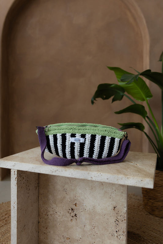 Gehäkelte Tasche in Bananenform in den Farben Schwarz, Weiß, Matcha-Grün und Violet mit verstellbarem Umhängegurt steht auf einem kleinen Tisch aus Stein in Beigetönen. Im Hintergrund eine braune Limewash-Wand mit einer Bogen-Nische. Auf dem Boden ein runder Teppich aus Jute, sowie eine Pflanze mit großen grünen Blättern in einem hellbraunen Topf, das Material ähnlich Bambus, die von rechts in das Bild ragt.