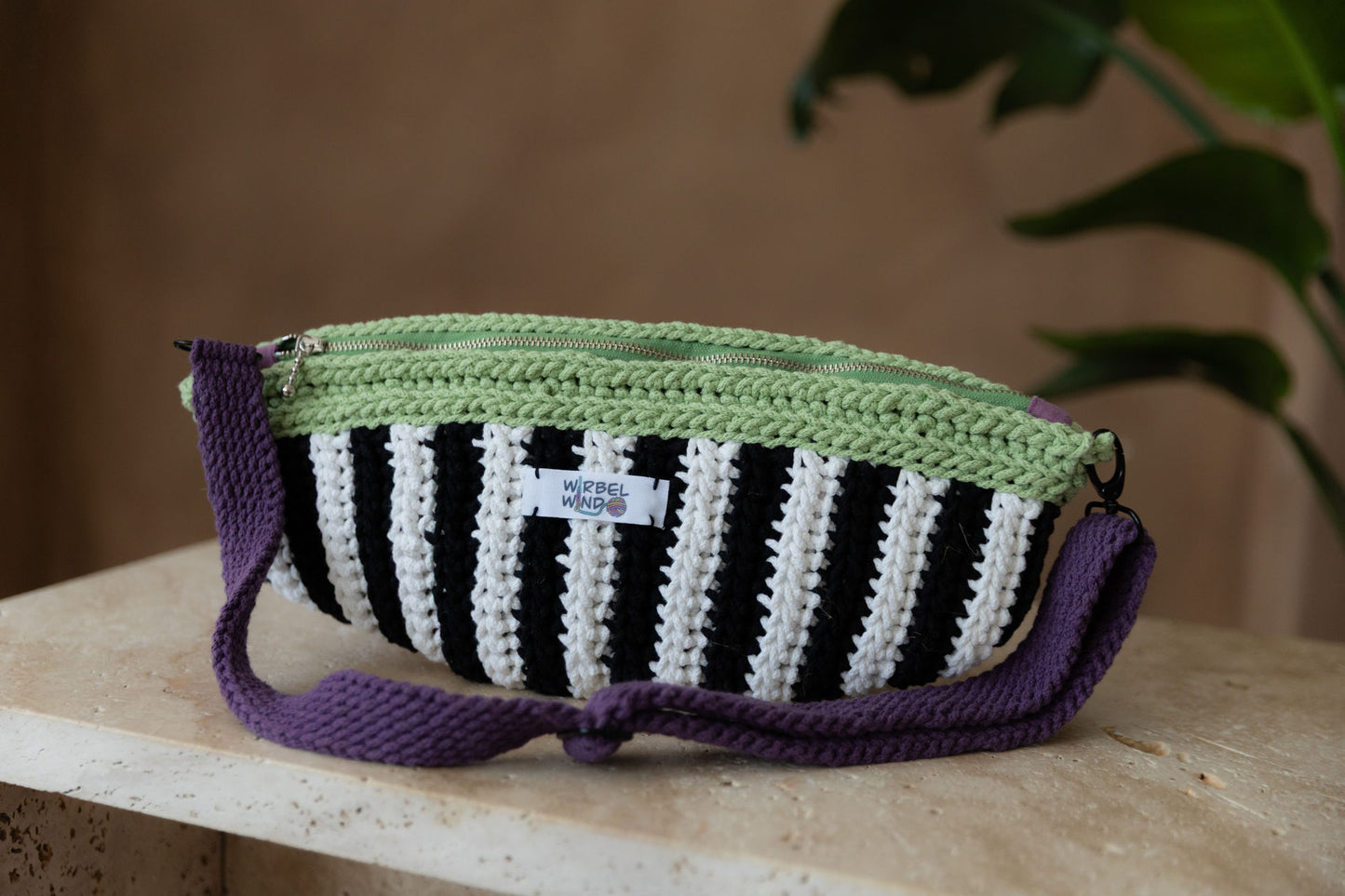 Gehäkelte Tasche in Bananenform in den Farben Schwarz, Weiß, Matcha-Grün und Violet mit verstellbarem Umhängegurt steht auf einem kleinen Tisch aus Stein in Beigetönen. Tasche füllt fast das ganze Bild aus, sehr stark vergrößert zum vorangegangenen Bild. Im Hintergrund sehr verschwommen die braune Wand und dei Pflanze mit großen grünen Blättern, die von rechts in das Bild ragt.