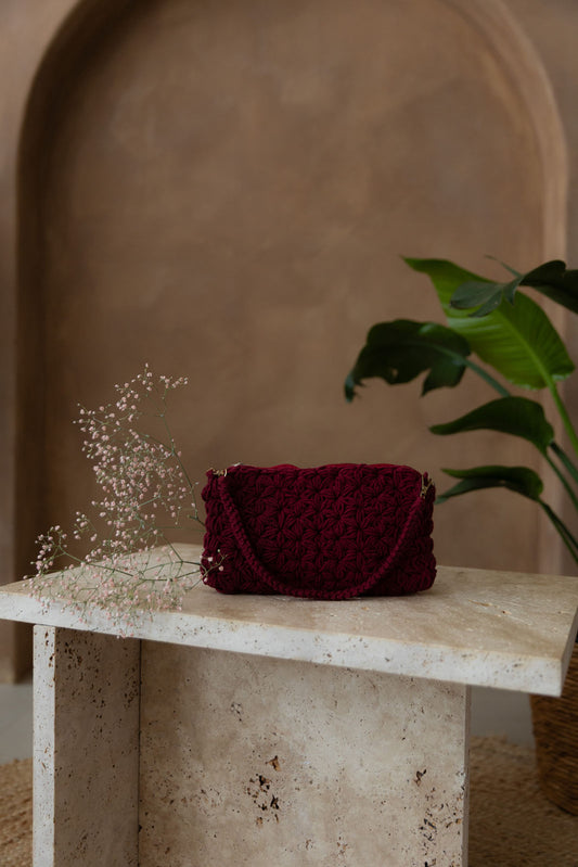 Gehäkelte Schultertasche in der Farbe Weinrot in rechteckiger Form mit nicht verstellbarem Schultergurt steht auf einem kleinen Tisch aus Stein in Beigetönen. Im Hintergrund eine braune Limewash-Wand mit einer Bogen-Nische. Auf dem Boden ein runder Teppich aus Jute, sowie eine Pflanze mit großen grünen Blättern, die von rechts in das Bild ragen, in einem hellbraunen Topf, das Material ähnlich Bambus. Auf der linken Seite der Tasche liegt ein Zweig rosaner Schleierkraut.