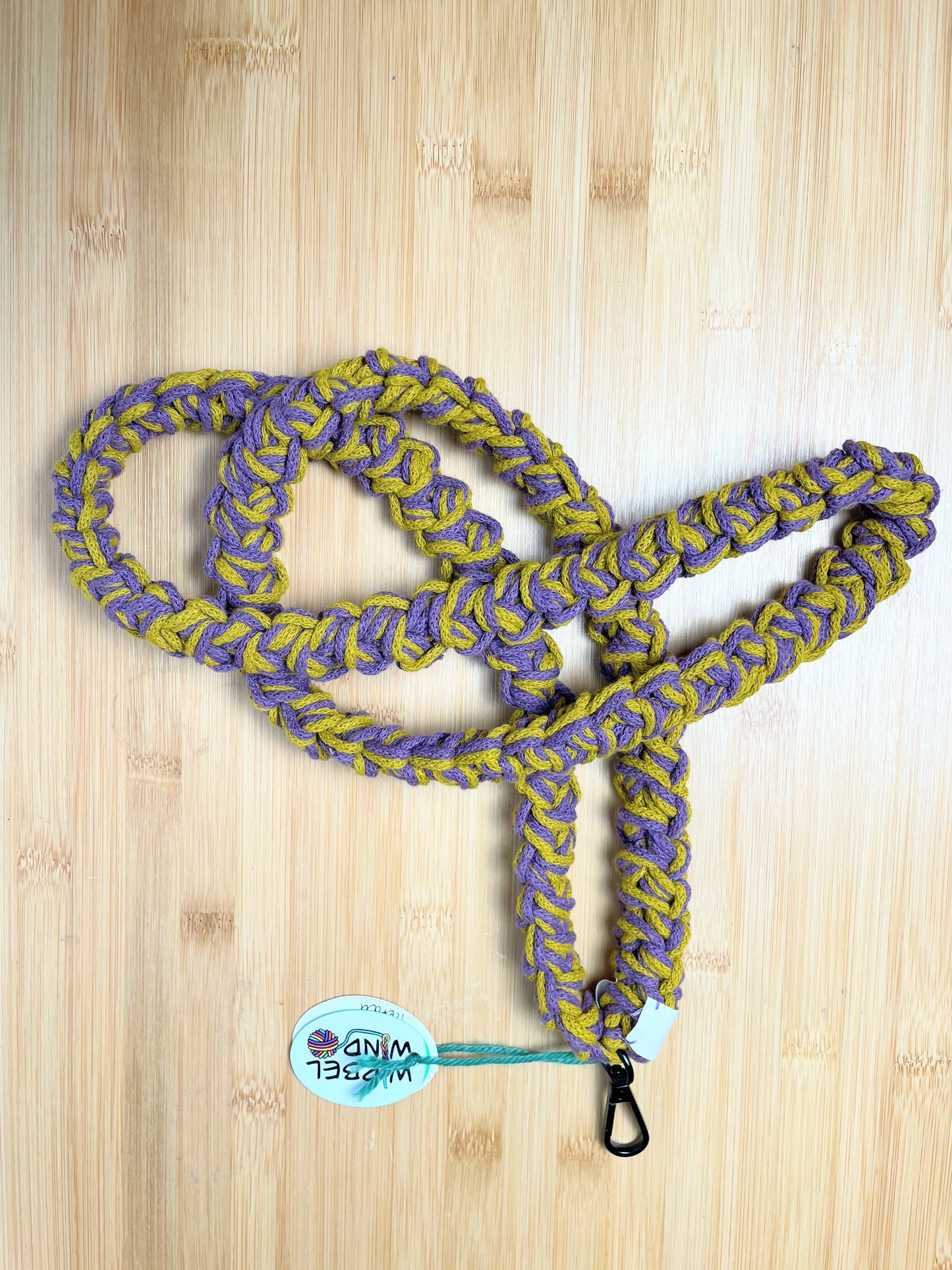 Gewürzgelb-Violet-meliertes Lanyard kringelt sich. Wirbelwindlogo am unteren Ende, direkt über schwarzem Karabiner angenäht.