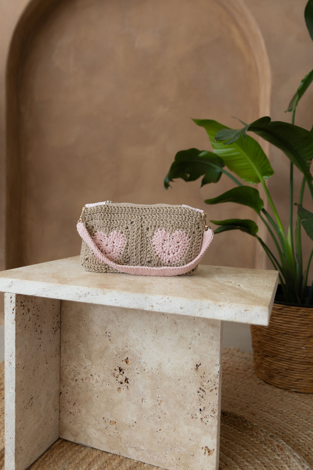 Gehäkelte rechteckige Schultertasche in Sandbraun mit vier Herzen in Pastellrosa, der nicht verstellbarem Schultergurt ist ebenfalls Hellrosa. Sie steht auf einem kleinen Tisch aus Stein in Beigetönen. Im Hintergrund eine braune Limewash-Wand mit einer Bogen-Nische. Auf dem Boden ein runder Teppich aus Jute, sowie eine Pflanze mit großen grünen Blättern, die von rechts in das Bild ragen, in einem hellbraunen Topf, das Material ähnlich Bambus.