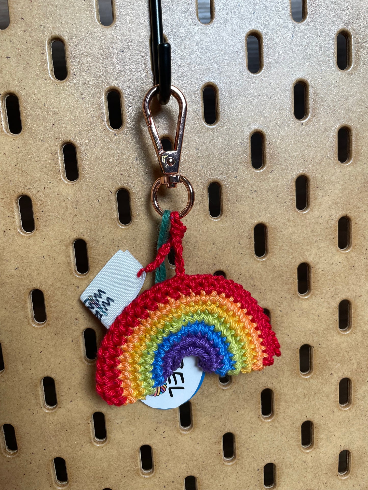 Gehäkelter Regenbogen Schlüsselanhänger mit Karabiner in den 6 Farben Regenbogens: Rot, Hellorange, Gelb, Hellgrün, Blau, Dunkellila. Außerdem hängt das Logo von Wirbelwind ebenfalls am Karabiner.