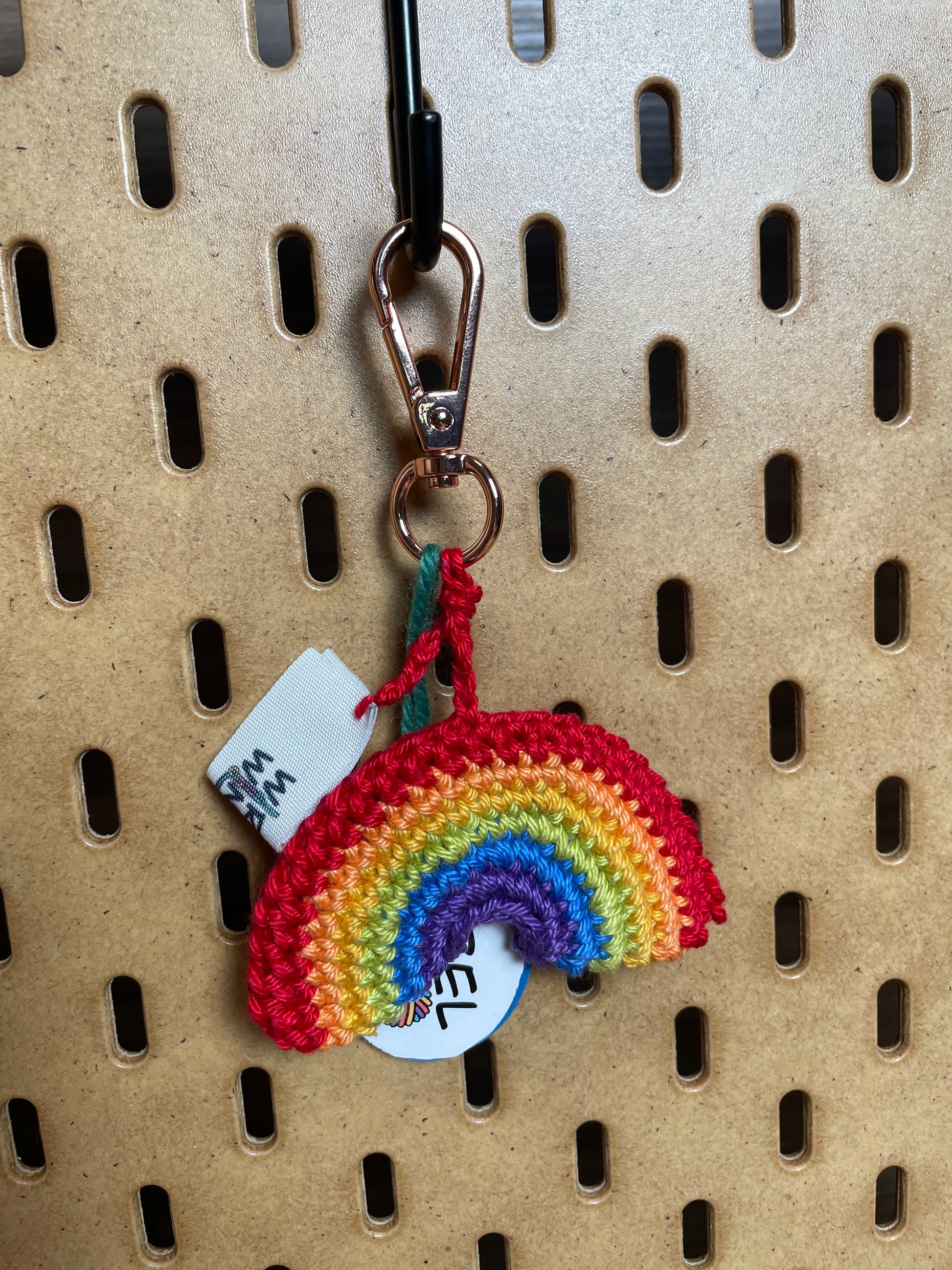 Gehäkelter Regenbogen Schlüsselanhänger mit Karabiner in den 6 Farben Regenbogens: Rot, Hellorange, Gelb, Hellgrün, Blau, Dunkellila. Außerdem hängt das Logo von Wirbelwind ebenfalls am Karabiner.