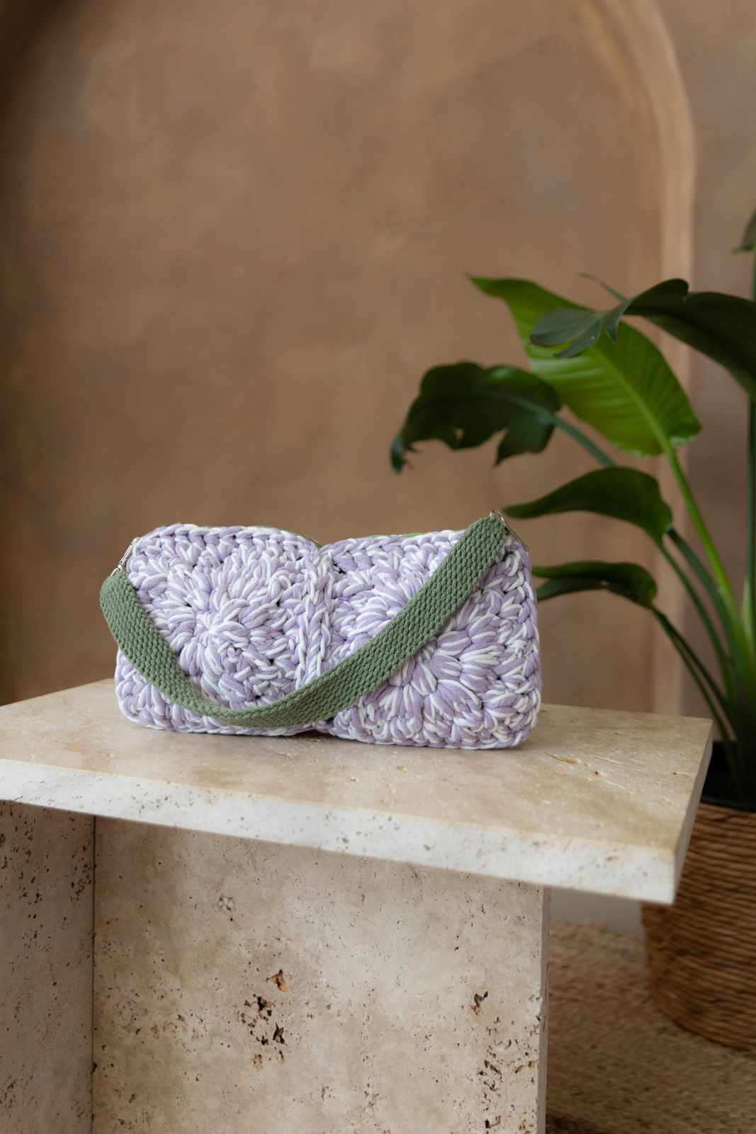 Gehäkelte rechteckige Schultertasche in den Farben Lavendel und Weiß, der nicht verstellbarem Schultergurt ist Eukalyptusgrün. Sie steht auf einem kleinen Tisch aus Stein in Beigetönen. Im Hintergrund eine braune Limewash-Wand mit einer Bogen-Nische. Auf dem Boden ein runder Teppich aus Jute, sowie eine Pflanze mit großen grünen Blättern, die von rechts in das Bild ragen, in einem hellbraunen Topf, das Material ähnlich Bambus. 