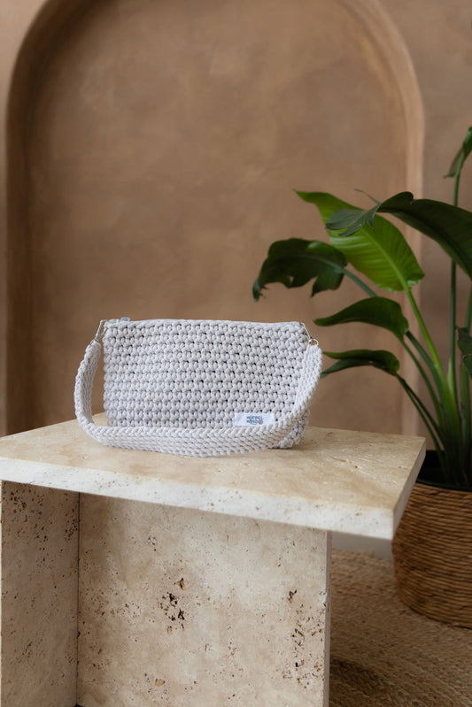 Gehäkelte rechteckige Schultertasche mit nicht verstellbarem Schultergurt in der Farbe Moonlight, ein fast weißes Helblau steht auf einem kleinen Tisch aus Stein in Beigetönen. Im Hintergrund eine braune Limewash-Wand mit einer Bogen-Nische. Auf dem Boden ein runder Teppich aus Jute, sowie eine Pflanze mit großen grünen Blättern, die von rechts in das Bild ragen, in einem hellbraunen Topf, das Material ähnlich Bambus.