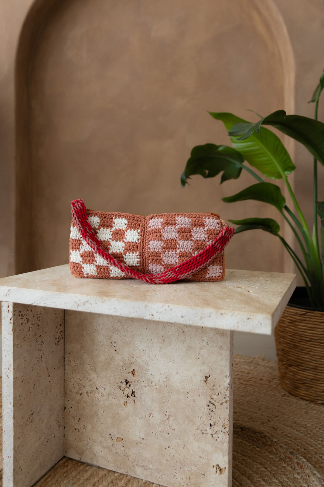 Gehäkelte rechteckige Schultertasche mit nicht verstellbarem Schultergurt und Karomuster steht auf einem kleinen Tisch aus Stein in Beigetönen. Im Hintergrund eine braune Limewash-Wand mit einer Bogen-Nische. Auf dem Boden ein runder Teppich aus Jute, sowie eine Pflanze mit großen grünen Blättern, die von rechts in das Bild ragen, in einem hellbraunen Topf, das Material ähnlich Bambus. Die terrakottafarbene Seite ist zu sehen.