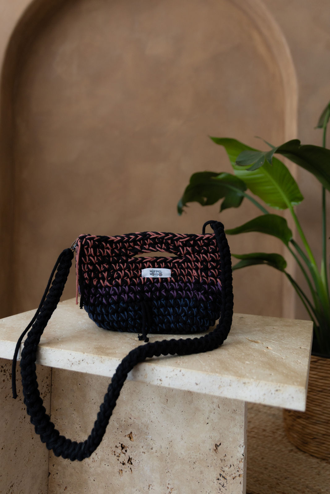 Gehäkelte viereckige Umhängetasche in den Farben Schwarz, Pink, Lila, Blau. Der nicht verstellbarem Schultergurt ist Schwarz und hängt vor der Tasche nach unten. Sie steht auf einem kleinen Tisch aus Stein in Beigetönen. Im Hintergrund eine braune Limewash-Wand mit einer Bogen-Nische. Auf dem Boden ein runder Teppich aus Jute, sowie eine Pflanze mit großen grünen Blättern, die von rechts in das Bild ragen, in einem hellbraunen Topf, das Material ähnlich Bambus. 