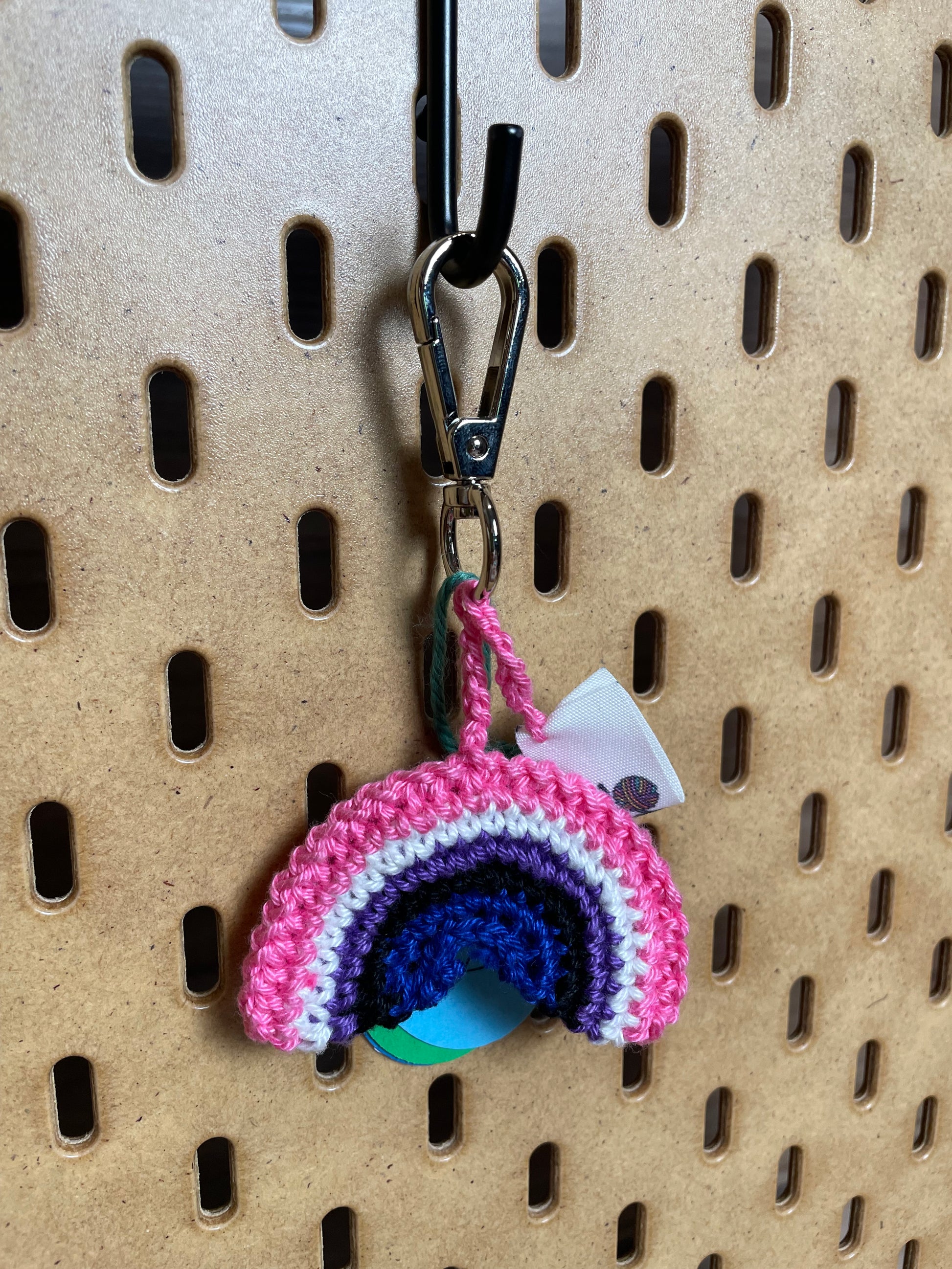 Gehäkelter Regenbogen Schlüsselanhänger mit Karabiner in den Farben der Genderfluid Pride Flagge: Pink, Weiß, Lila, Schwarz, Blau. Außerdem hängt das Logo von Wirbelwind ebenfalls am Karabiner.