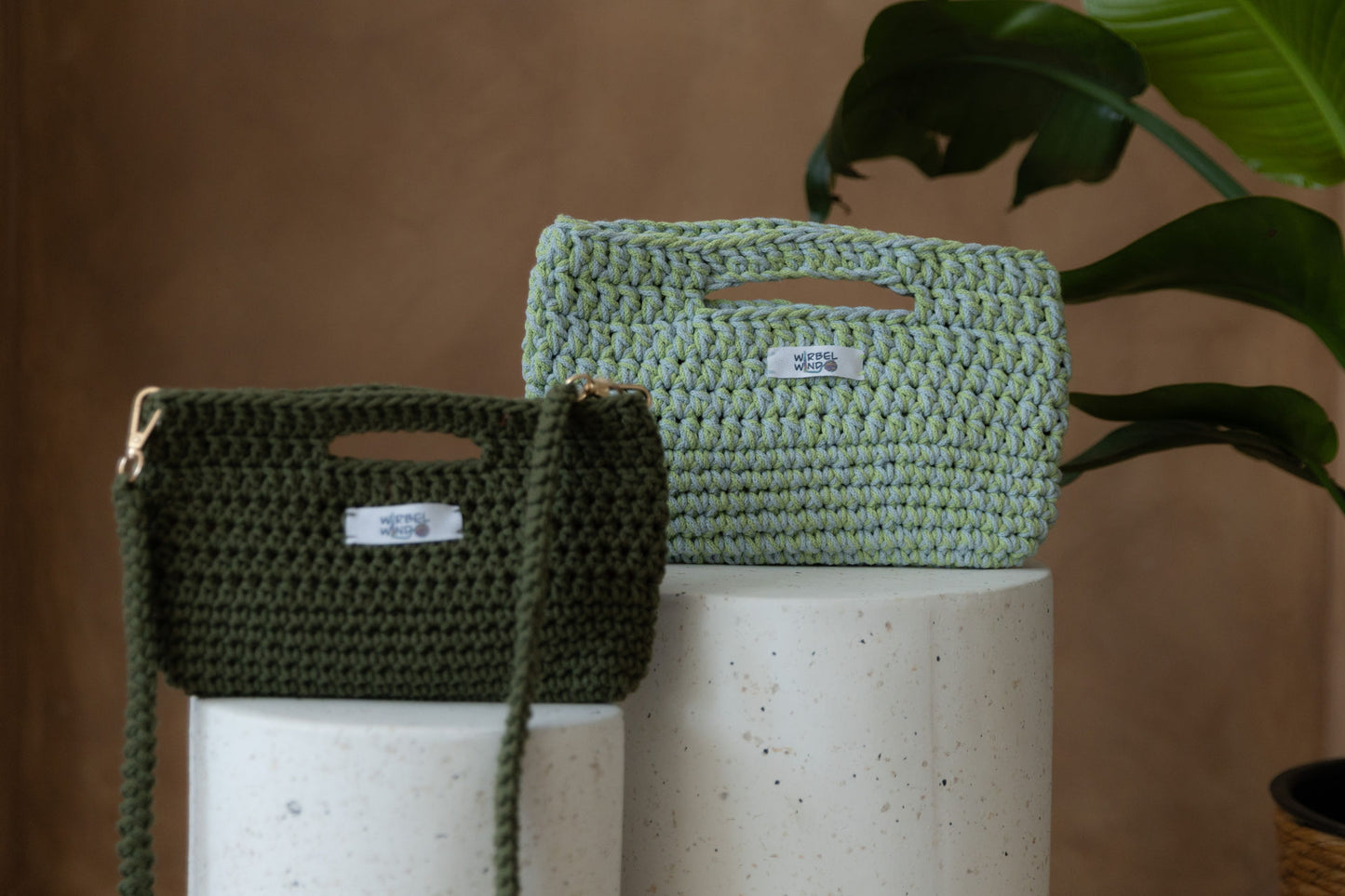Nahaufnahme der Molly Medium Tasche in Hellblau & Hellgrün und der Molly Mini Tasche in Dunkelgrün, wie sie auf den Bettonsäulen stehen. Sie füllen fast den ganzen Bildschirm aus. Die kleine Tasche im Vordergrund ist unscharf, der Fokus liegt aus der hinteren.