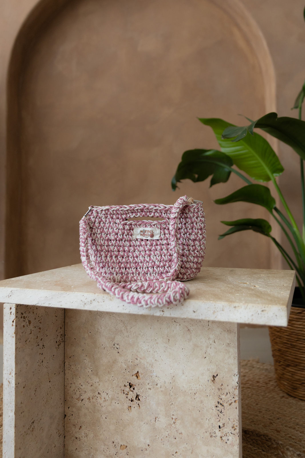 Gehäkelte viereckige Umhängetasche in Nude-Weiß und Blossom-Pink verwoben. Der Schultergurt hast dieselben Farben und hängt vor der Tasche herunter und kringelt sich auf dem Tisch. Dieser ist aus Stein in Beigetönen. Im Hintergrund eine braune Limewash-Wand mit einer Bogen-Nische. Auf dem Boden ein runder Teppich aus Jute, sowie eine Pflanze mit großen grünen Blättern, die von rechts in das Bild ragen, in einem hellbraunen Topf, das Material ähnlich Bambus. 