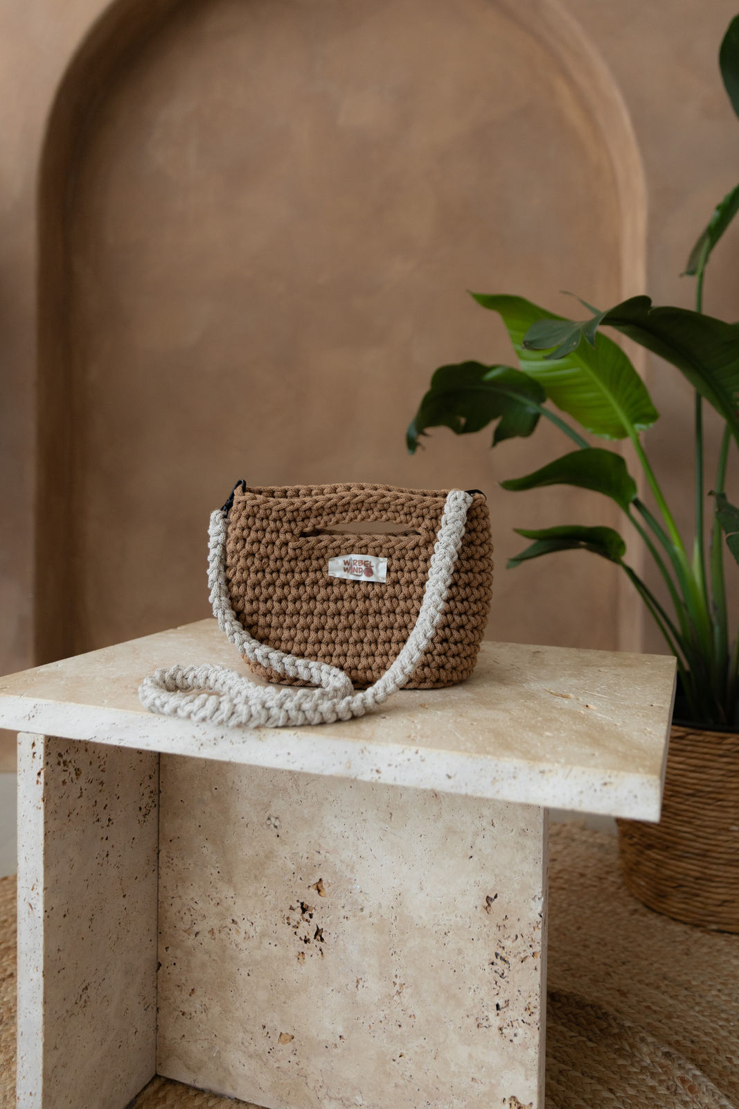 Gehäkelte viereckige Umhängetasche in Karamellbraun. Der Schultergurt ist Beige mit kleinen Sprenkeln aus Schwarz und Karamell und hängt vor der Tasche herunter und kringelt sich auf dem Tisch. Dieser ist aus Stein in Beigetönen. Im Hintergrund eine braune Limewash-Wand mit einer Bogen-Nische. Auf dem Boden ein runder Teppich aus Jute, sowie eine Pflanze mit großen grünen Blättern, die von rechts in das Bild ragen, in einem hellbraunen Topf, das Material ähnlich Bambus. 