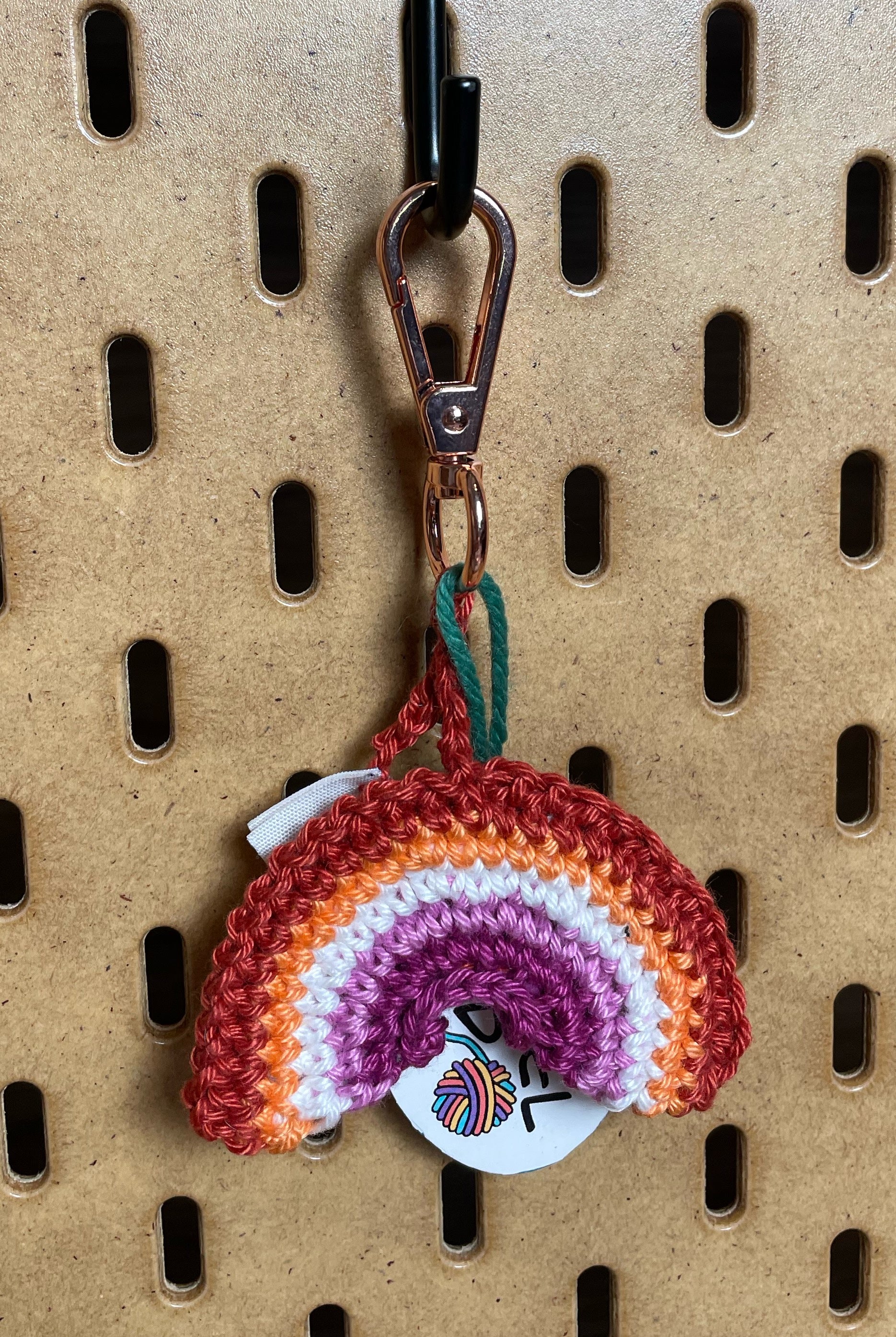 Gehäkelter Regenbogen Schlüsselanhänger mit Karabiner in den 5 Farben der Lesbischen Pride Flagge: Orangerot, Hellorange, Weiß, Hellpink, Dunkelpink. Außerdem hängt das Logo von Wirbelwind ebenfalls am Karabiner.