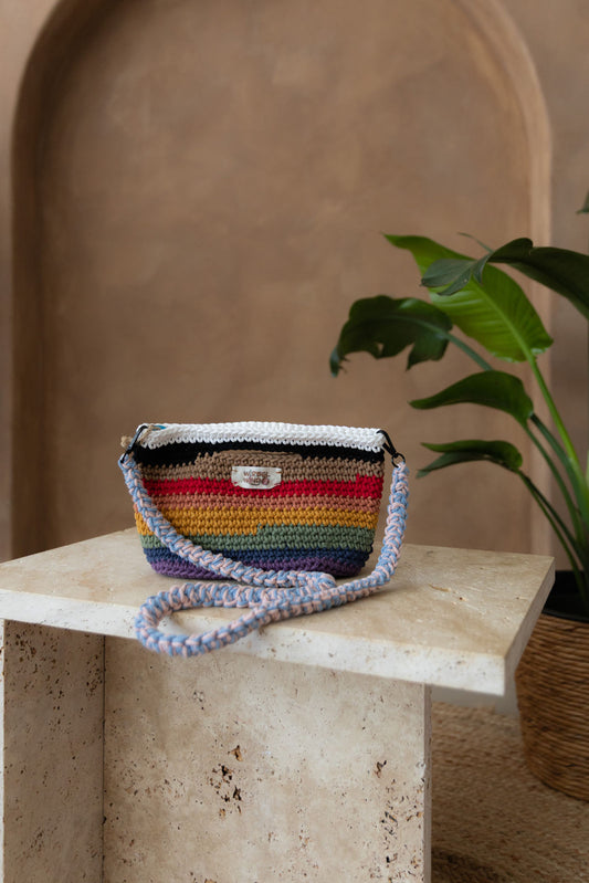 Gehäkelte viereckige Umhängetasche in den Farben des Regenbogens. Der Schultergurt hängt vor der Tasche nach unten. Sie steht auf einem kleinen Tisch aus Stein in Beigetönen. Im Hintergrund eine braune Limewash-Wand mit einer Bogen-Nische. Auf dem Boden ein runder Teppich aus Jute, sowie eine Pflanze mit großen grünen Blättern, die von rechts in das Bild ragen, in einem hellbraunen Topf, das Material ähnlich Bambus. 