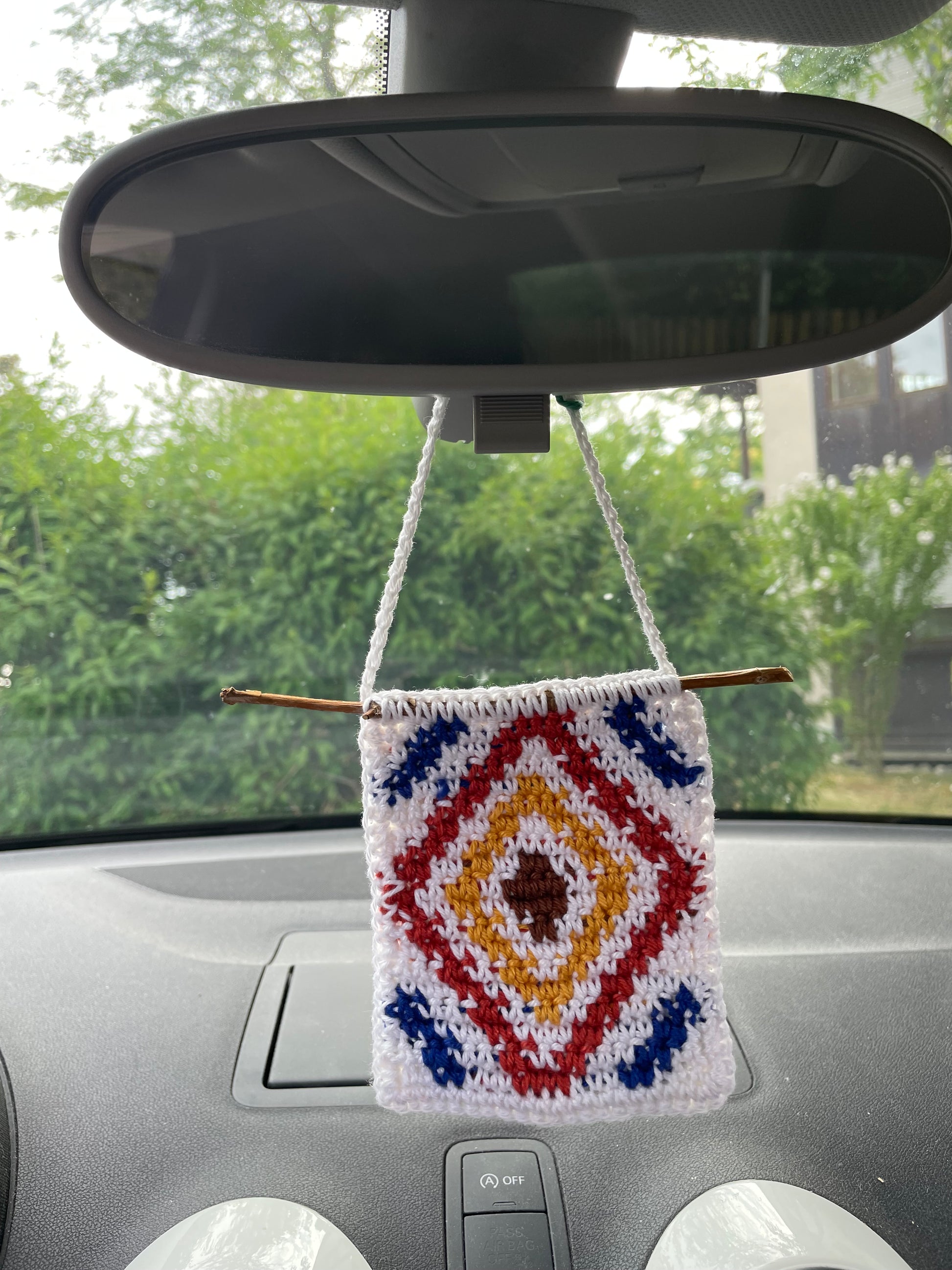 Gehäkelter Anhänger mit geometrischem Muster hängt in Auto an Rückspiegel. Mitte braune Raute, umrandet von weißer, umrandet von rot/orangener, umrandet von weißer Raute. Nachfolgende blaue Raute ragt an Ecken aus Bild heraus, ebenso folgende weiße Raute.