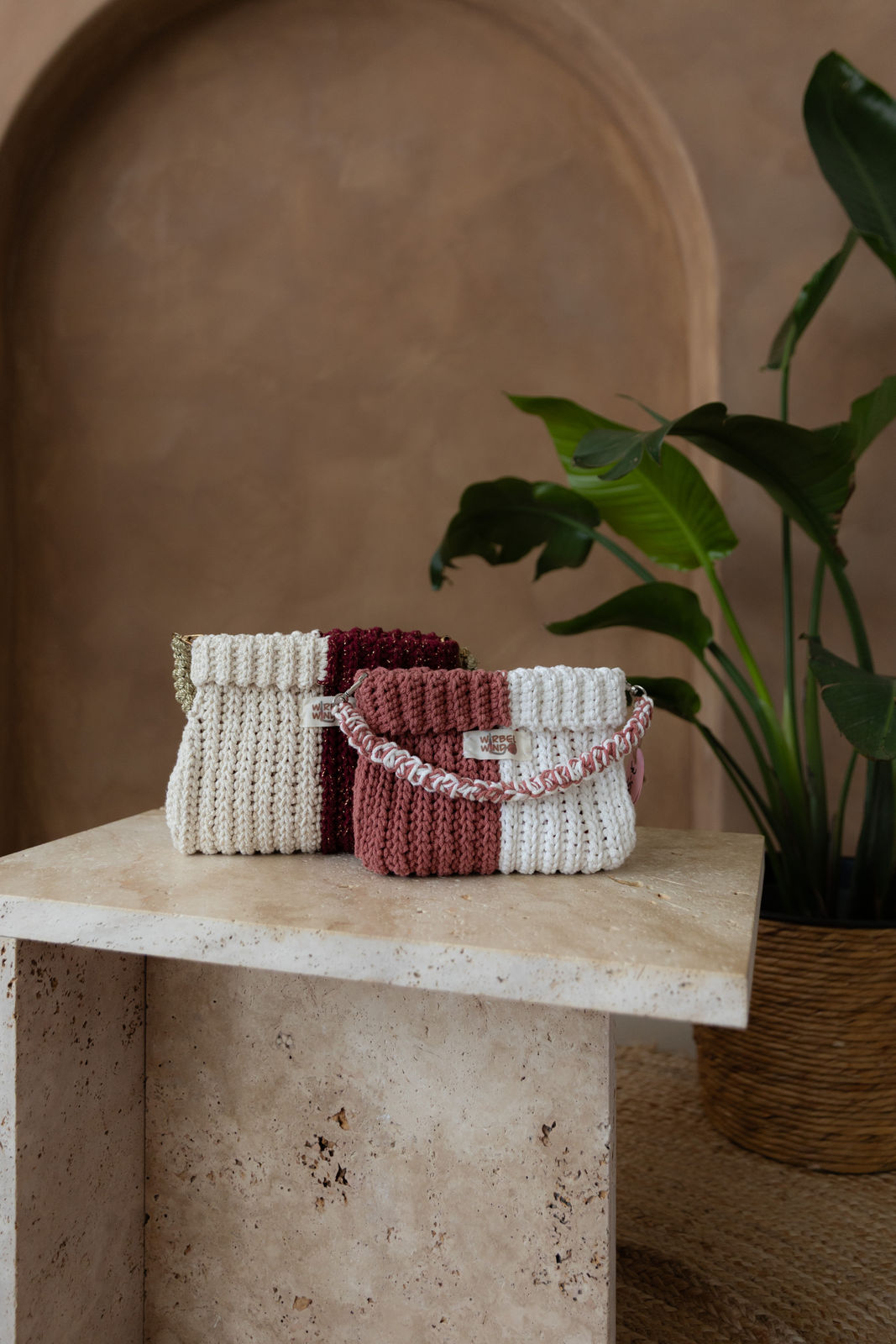 2 Gabbie Taschen stehen auf Steintisch voreinander. Vorne die etw. kleinere SAKURA Edition, dahinter die dunklere Tasche der ROMANTIK Kollektion. Hintergrund ist braune lime-wash Wand mit Bogen. Rechts ragt große grüne Pflanze ins Bild.