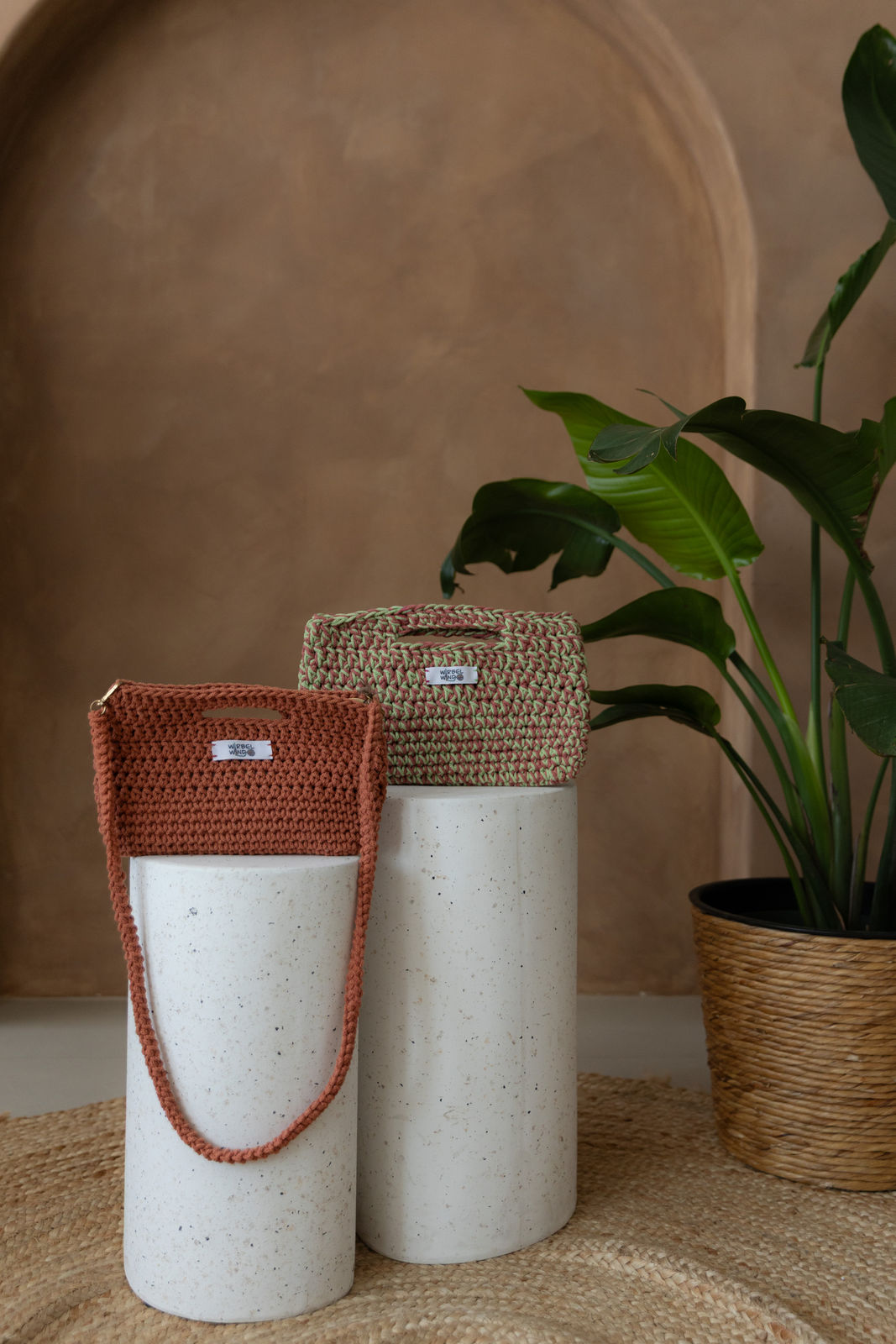 2 unterschiedlich hohe runde Betonsäulen. Auf ihnen rechts und etwas nach hinten versetzt eine Molly Medium Tasche in Pink & Hellgrün, davor und links eine Molly Mini Tasche in Orange, ihr Umhängegurt hängt vorne nach unten. Rechts ragt grüne Pflanze ins Bild. Hintergrund ist braune Limewash Wand mit Bogen.