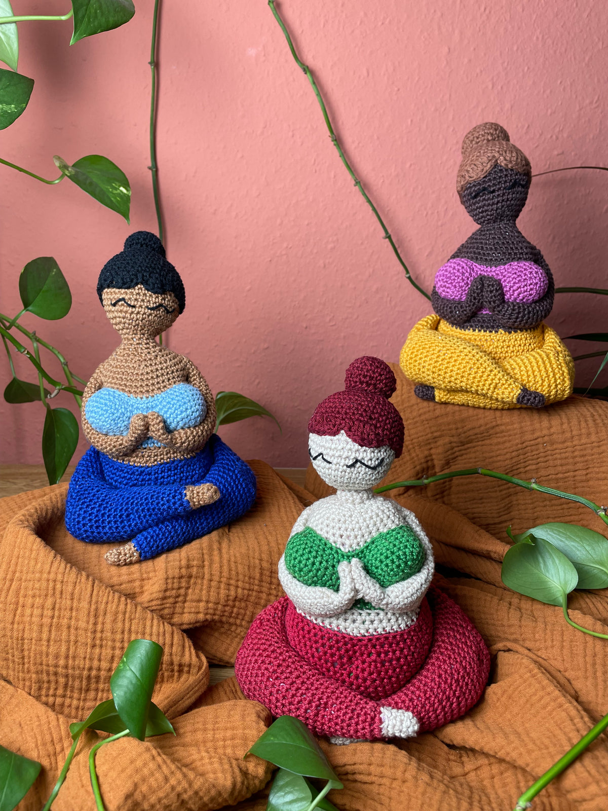 Gehäkelte Yoga Figuren im Schneidersitz, Hände vor dem Herzen zusammengeführt, verschiedene Haut- und Haarfarben. Hintergrund terrakottafarben, Efeututenranke verläuft durchs Bild.