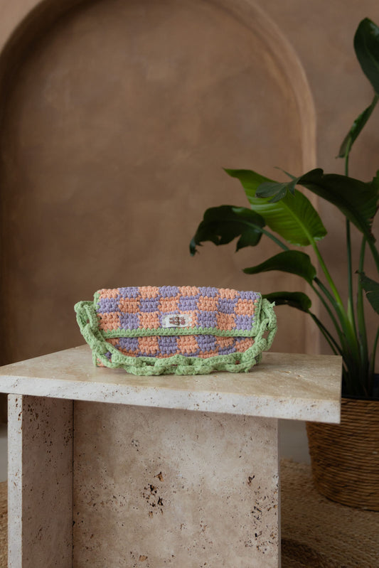 Gehäkelte Tasche in pfirsich-lavendel-kariert, umrisse und Schultergurt Matchagrün steht auf einem kleinen Tisch aus Stein in Beigetönen. Im Hintergrund eine braune Limewash-Wand mit einer Bogen-Nische. Auf dem Boden ein runder Teppich aus Jute, sowie eine Pflanze mit großen grünen Blättern in einem hellbraunen Topf, das Material ähnlich Bambus.