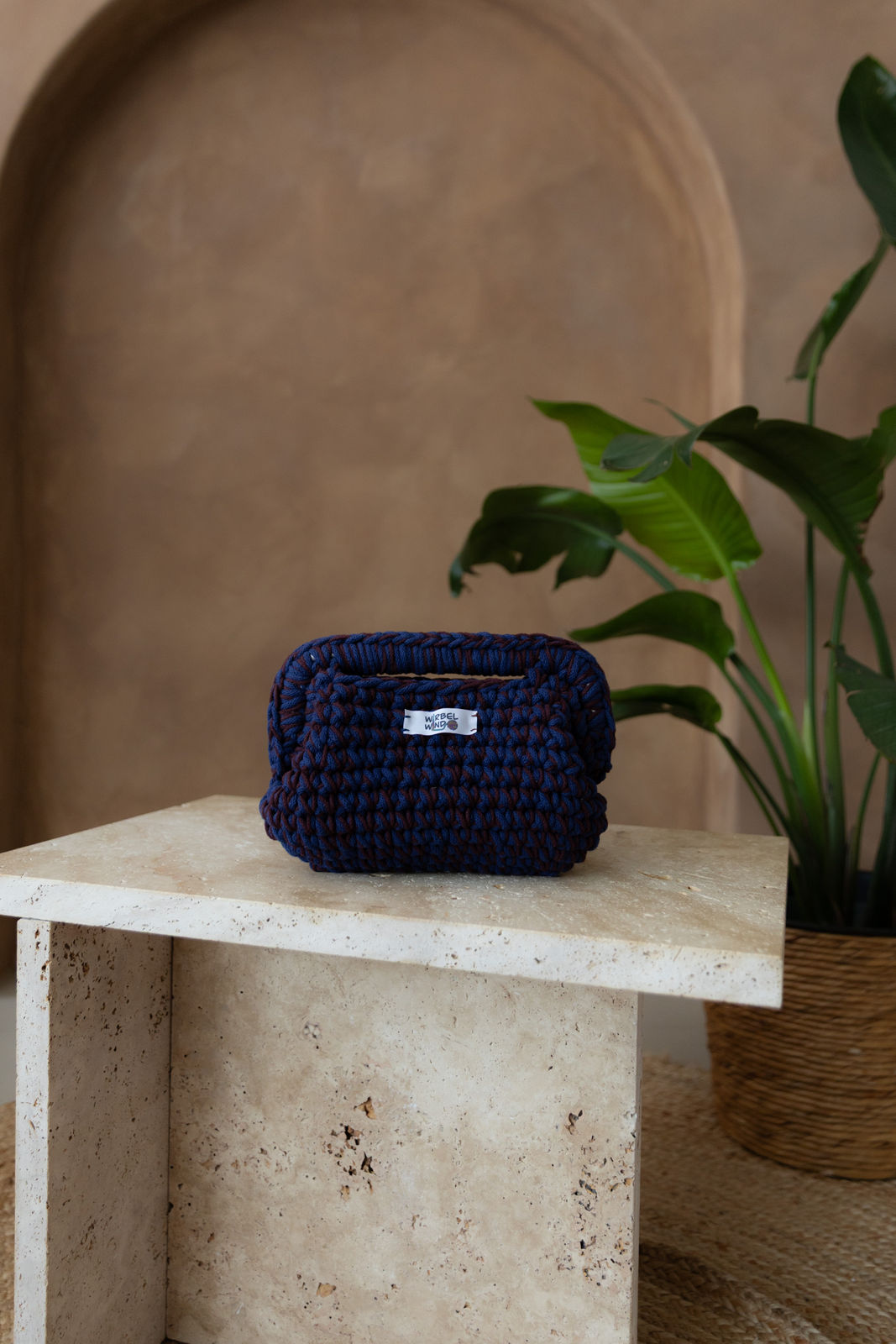 Gehäkelte, bauchige Clutch mit Obergriff in den Farben Burgunder-Rot und Tinten-Blau steht auf einem kleinen Tisch aus Stein in Beigetönen. Im Hintergrund eine braune Limewash-Wand mit einer Bogen-Nische. Auf dem Boden ein runder Teppich aus Jute, sowie eine Pflanze mit großen grünen Blättern in einem hellbraunen Topf, das Material ähnlich Bambus.