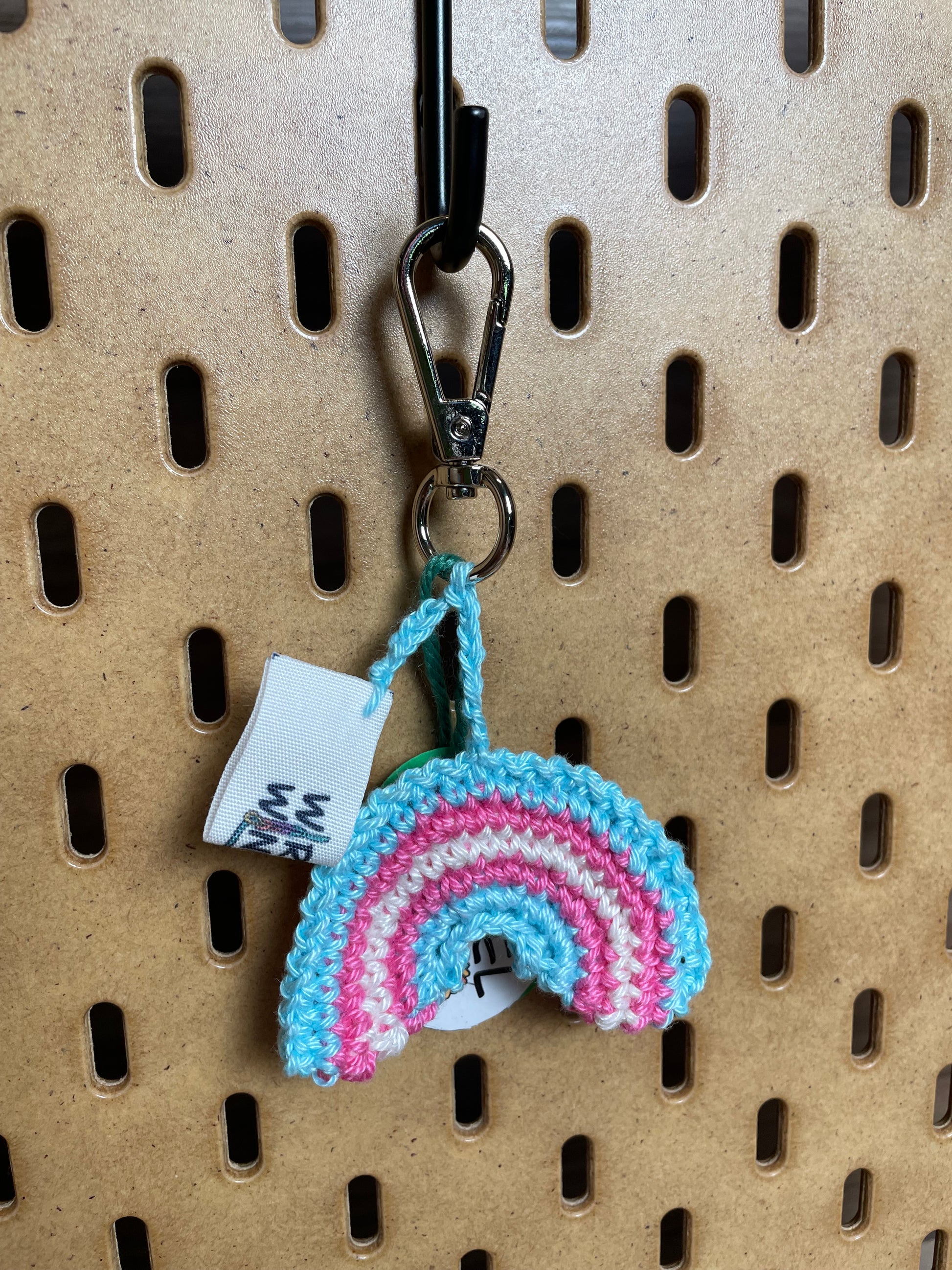 Gehäkelter Regenbogen Schlüsselanhänger mit Karabiner in den Farben der Trans Pride Flagge: Hellblau, Hellpink, Weiß, Hellpink, Hellblau. Außerdem hängt das Logo von Wirbelwind ebenfalls am Karabiner.