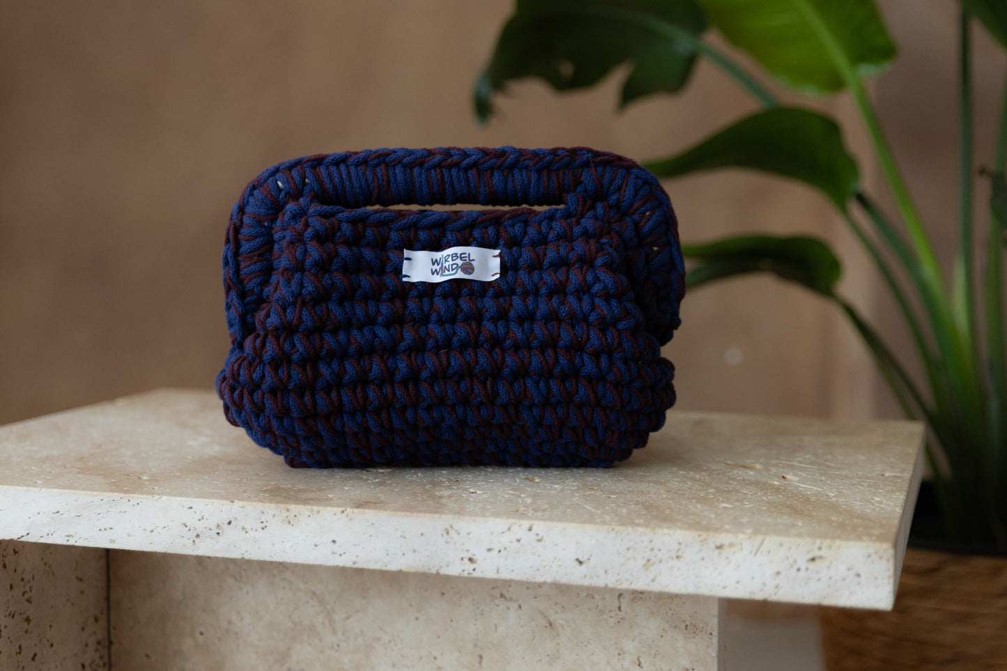 Gehäkelte, bauchige Clutch mit Obergriff in den Farben Burgunder-Rot und Tinten-Blau steht auf einem kleinen Tisch aus Stein in Beigetönen. Im Hintergrund eine braune Limewash-Wand mit einer Bogen-Nische. Auf dem Boden ein runder Teppich aus Jute, sowie eine Pflanze mit großen grünen Blättern in einem hellbraunen Topf, das Material ähnlich Bambus. Gleich dem vorherigen Foto, nur eine nähere Aufnahme.