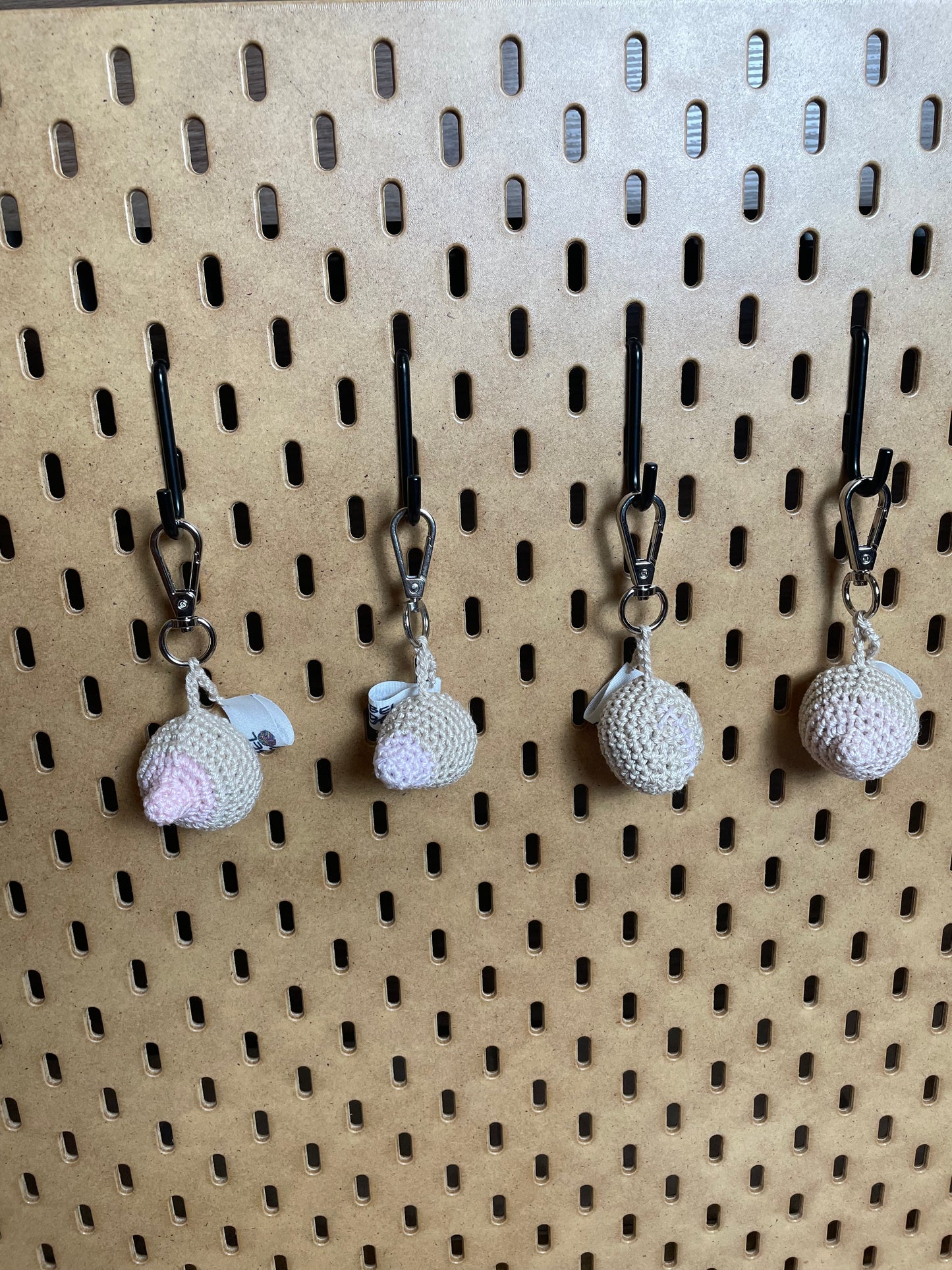 Frontale Aufnahme: 4 gehäkelte Brüste in Hellbeige mit Rosa Nippeln/Narben an silbernen Karabinern aufgehängt.