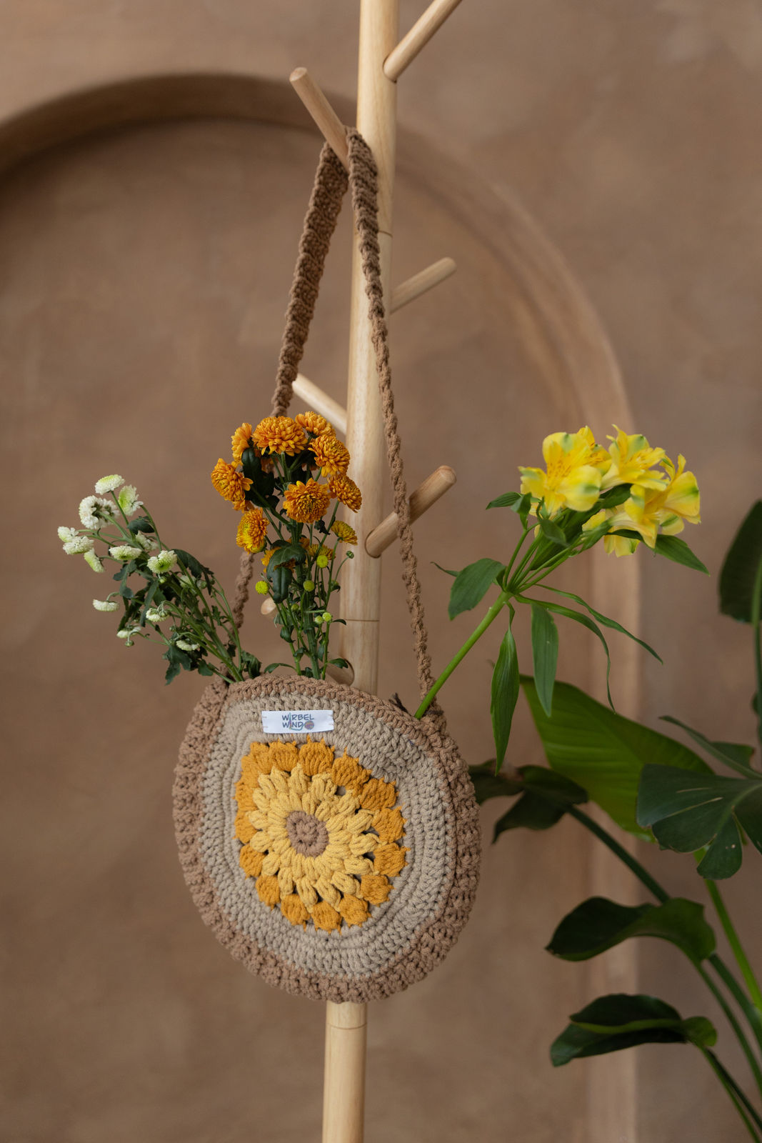 Gehäkelte runde Umhängetasche hängt an einem Garderobenständer aus Holz. Aus der Tasche ragen drei Blumen; eine gelb, eine orange und eine weiß. Alle Blumen haben mehrere Blüten. Im Hintergrund eine braune Limewash-Wand mit einer Bogen-Nische. Auf dem Boden am rechten Rand steht eine Pflanze mit großen grünen Blättern, die in das Bild ragen.