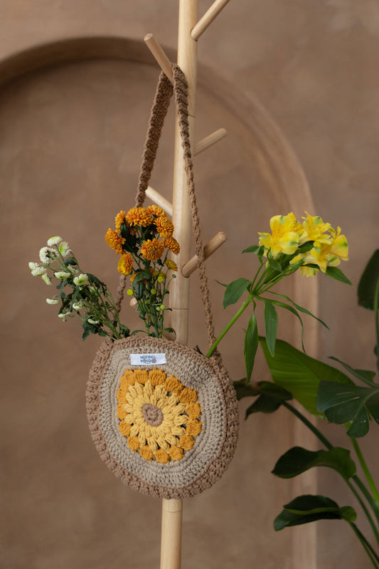 Gehäkelte runde Umhängetasche hängt an einem Garderobenständer aus Holz. Aus der Tasche ragen drei Blumen; eine gelb, eine orange und eine weiß. Alle Blumen haben mehrere Blüten. Im Hintergrund eine braune Limewash-Wand mit einer Bogen-Nische. Auf dem Boden am rechten Rand steht eine Pflanze mit großen grünen Blättern, die in das Bild ragen.