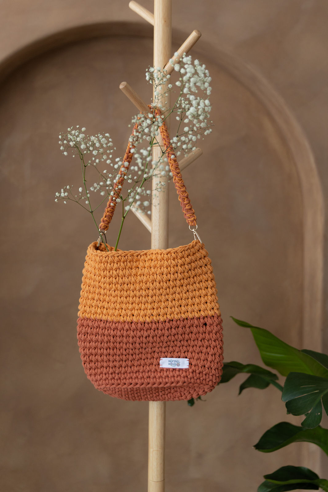 Gehäkelte Tasche, höher als breit, untere Hälfte Terrakotta-Orange, obere Hälfte helleres Orange, Henkel beide Farben verflochten, Karabiner des Henkels golden. In der Tasche steckt weißes Schleierkraut. Sie hängt an hölzernem Garderobenständer.