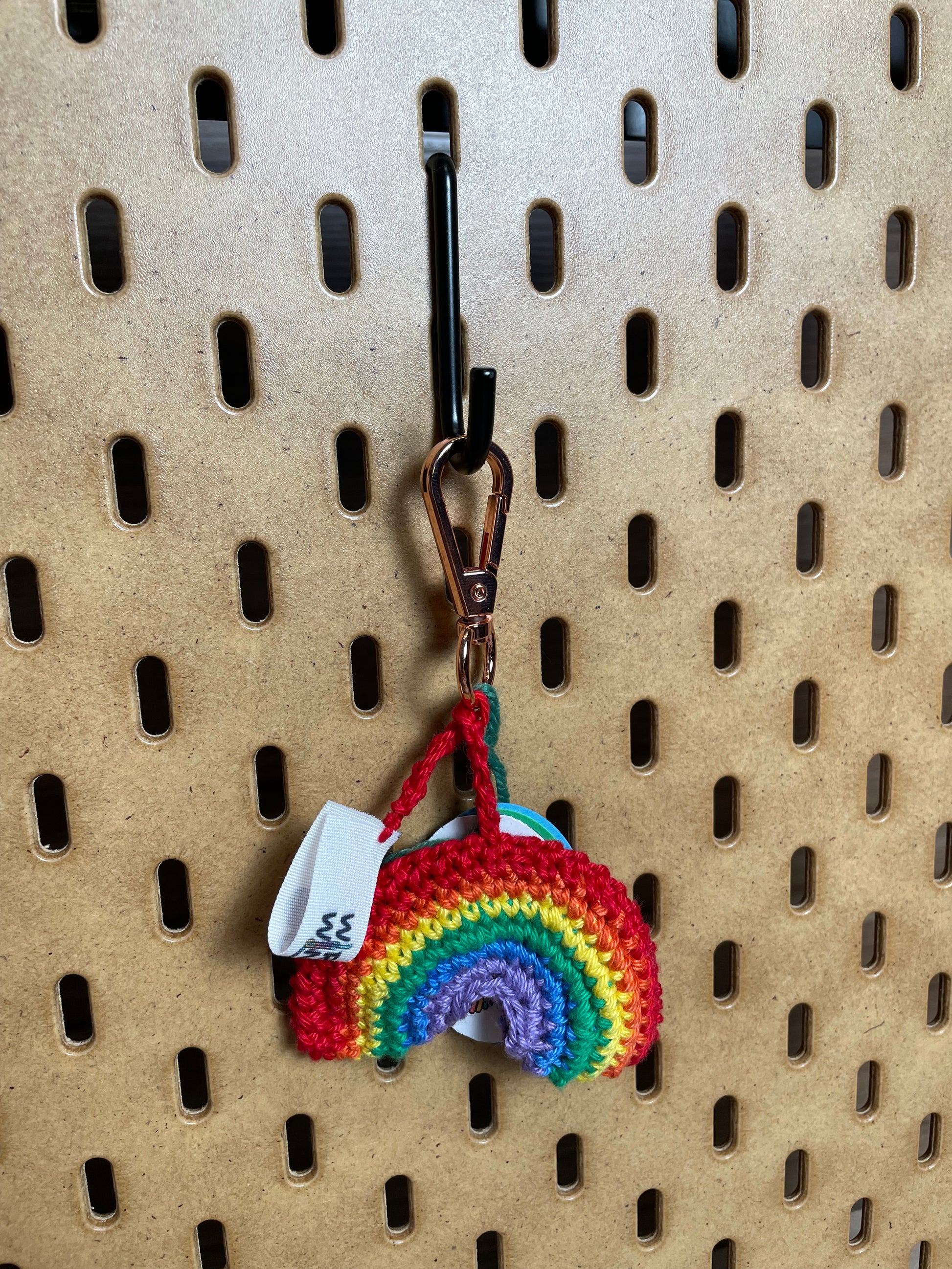 Gehäkelter Regenbogen Schlüsselanhänger mit Karabiner in den 6 Farben des Regenbogens: Rot, Orange, etwas helleres Gelb, etwas helleres Grün, Blau, Lila. Außerdem hängt das Logo von Wirbelwind ebenfalls am Karabiner.