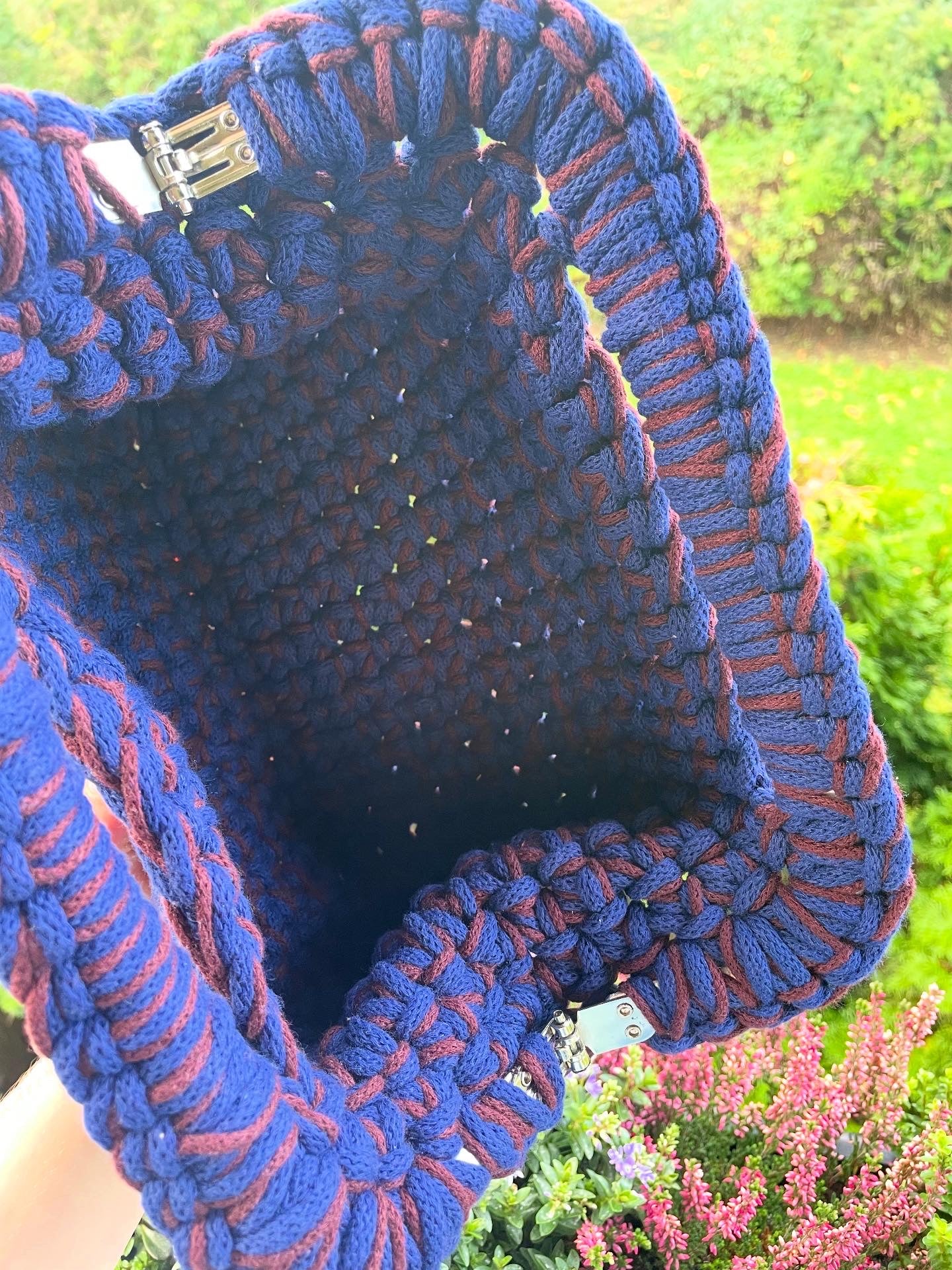 Aus dem linken Bildrand ragt ein Arm, der die Tasche am Boden über die Brüstung eines Balkons hält. Sie ist geöffnet und wir sehen, wie geräumig sie ist. Im Hintergrund eine sonnenbeschienene Wiese und Blumen.