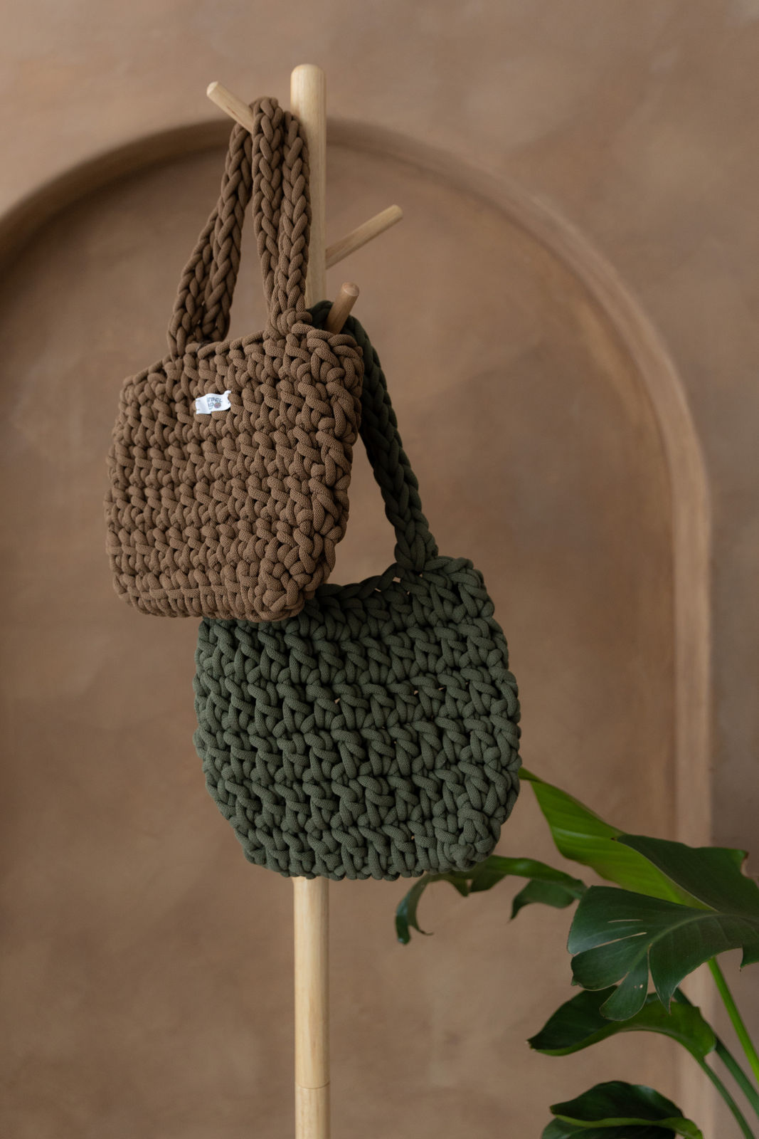 2 gehäkelte mittelgroße Schultertaschen in den Farben Nussbraun und Avokadogrün hängen an einem Garderobenständer aus Holz. Im Hintergrund eine braune Limewash-Wand mit einer Bogen-Nische. Am unteren rechten Rand ragen die großen grünen Blättern eienr Pflanze ins Bild.